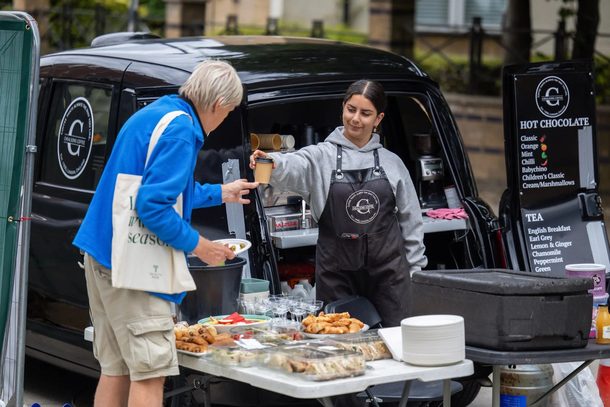 A food stall set up in the back of a black van selling baked goods, pancakes, and snacks at an outdoor event. A young woman with dark hair is handing a cup to an older man with short light-colored hair.