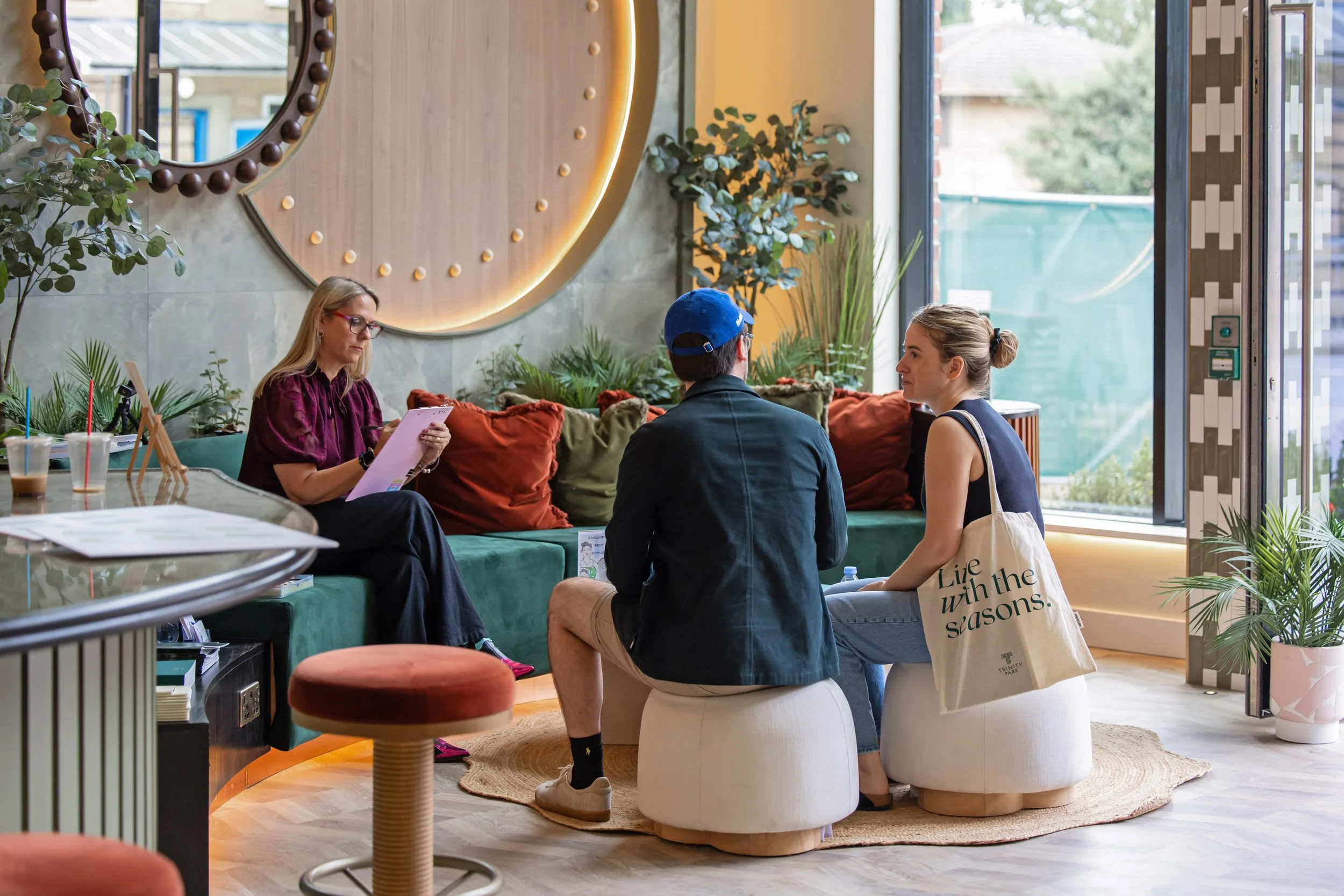 Three people seated on a green cushioned bench inside a modern cafe, with two women and a man. The woman on the left is reading a menu, the man and woman on the right are engaged in conversation. The cafe has plants, a large circular mirror with lights, and a glass door letting in natural light.