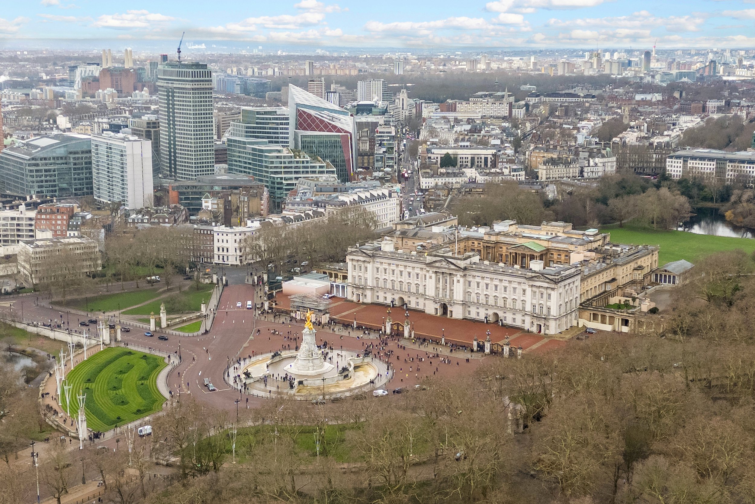 A panoramic view of a city skyline featuring modern skyscrapers and historic buildings. In the foreground is a large white building with classical architecture, a monument with statues, and a green park area with pathways and trees. The sky is partly cloudy.