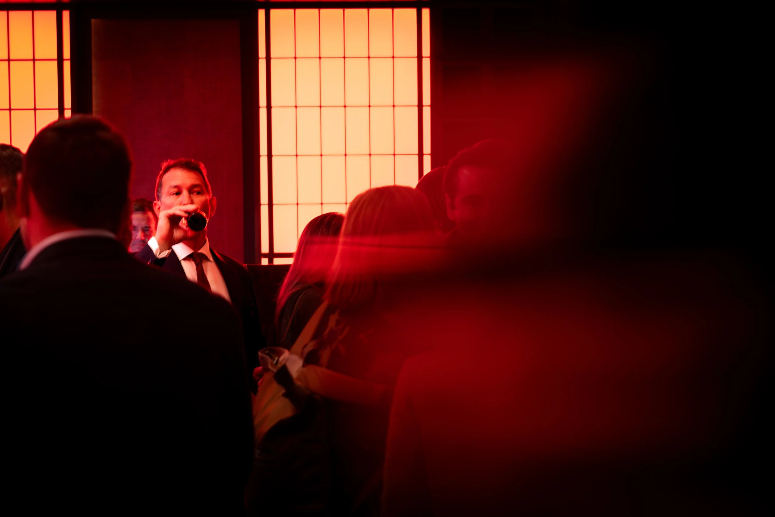 People at a social event with red lighting and Japanese-style shoji screens in the background, including a man in a suit drinking from a bottle.