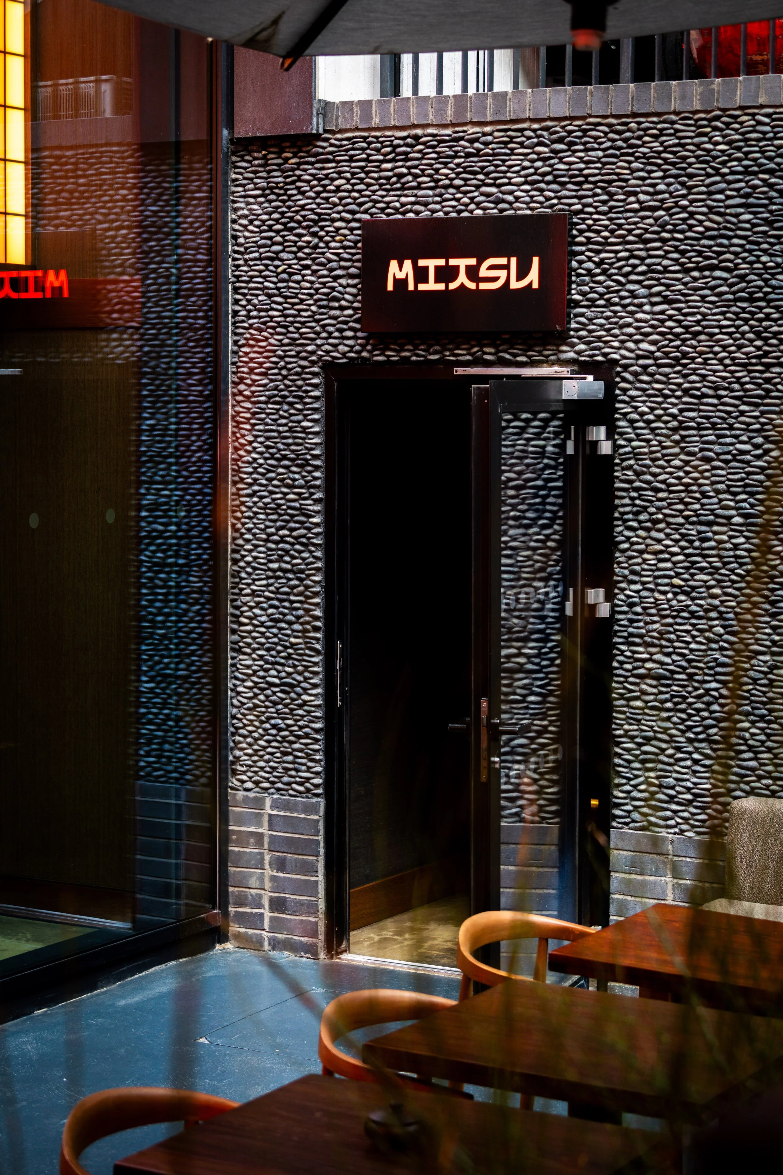 Interior of a restaurant with dark stone wall, doorway with glass door, tables, and wooden chairs.
