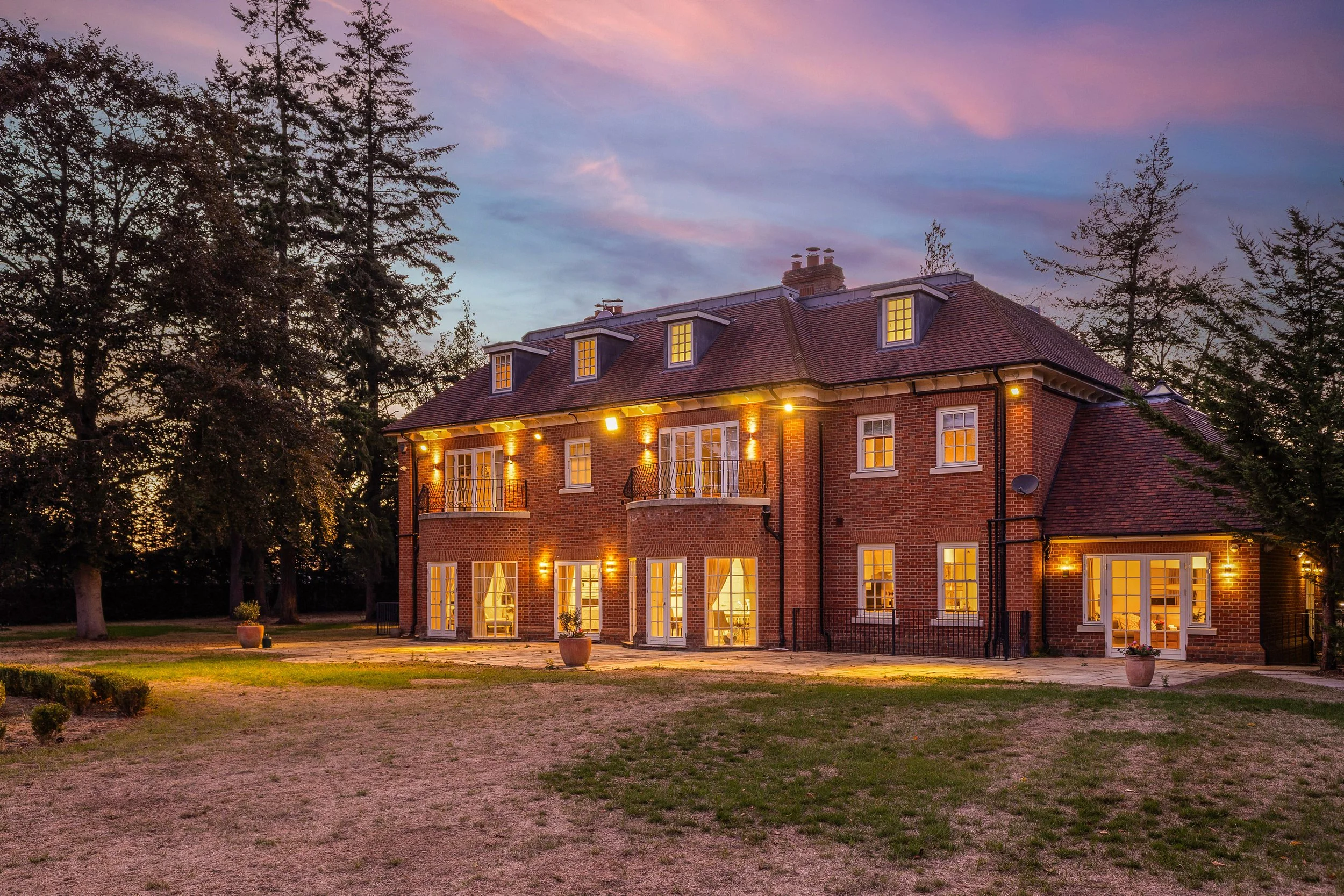 A large brick house with multiple lit windows, surrounded by trees at dusk with a colorful sky.
