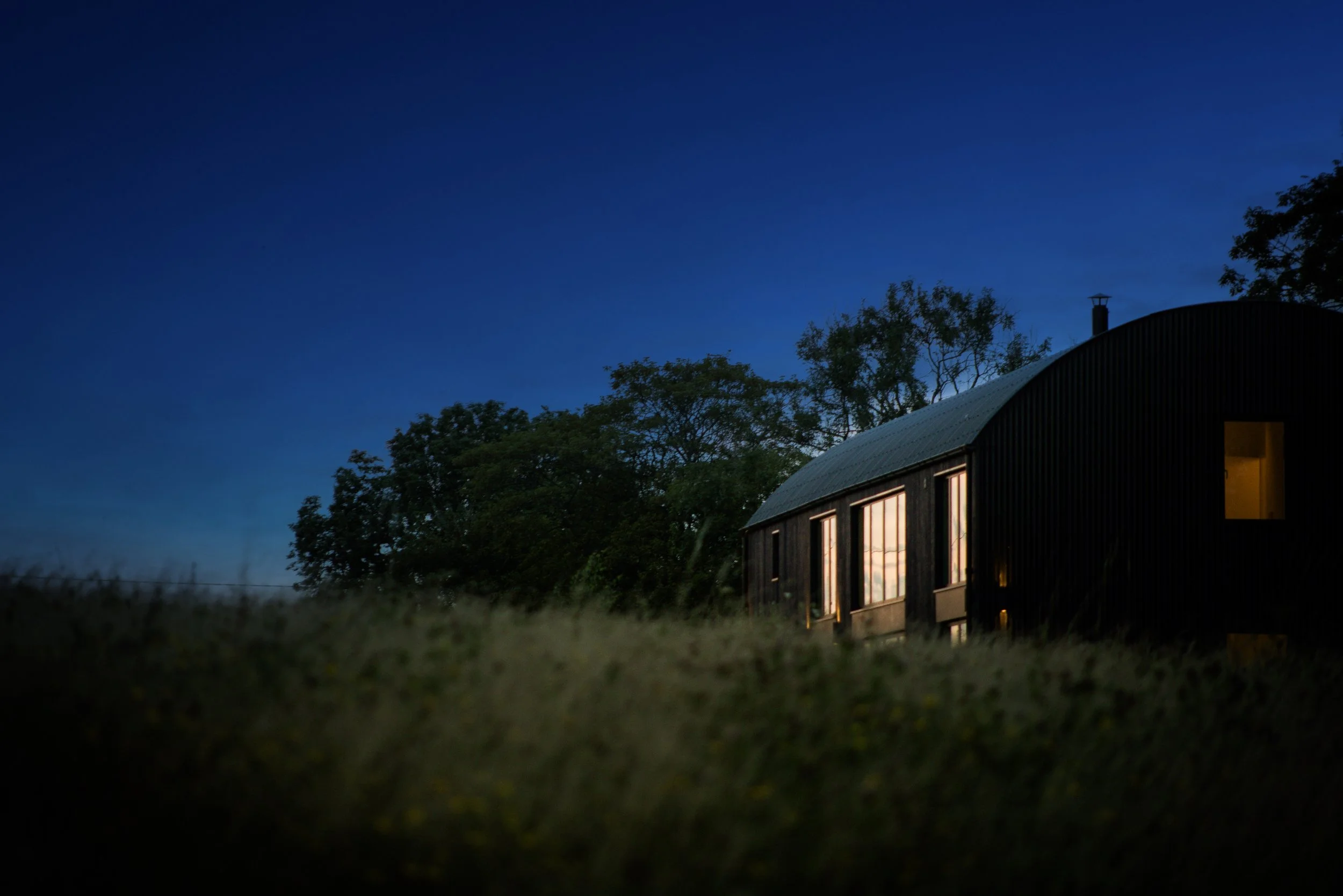 A house with illuminated windows at dusk, surrounded by trees, with a clear blue sky overhead.
