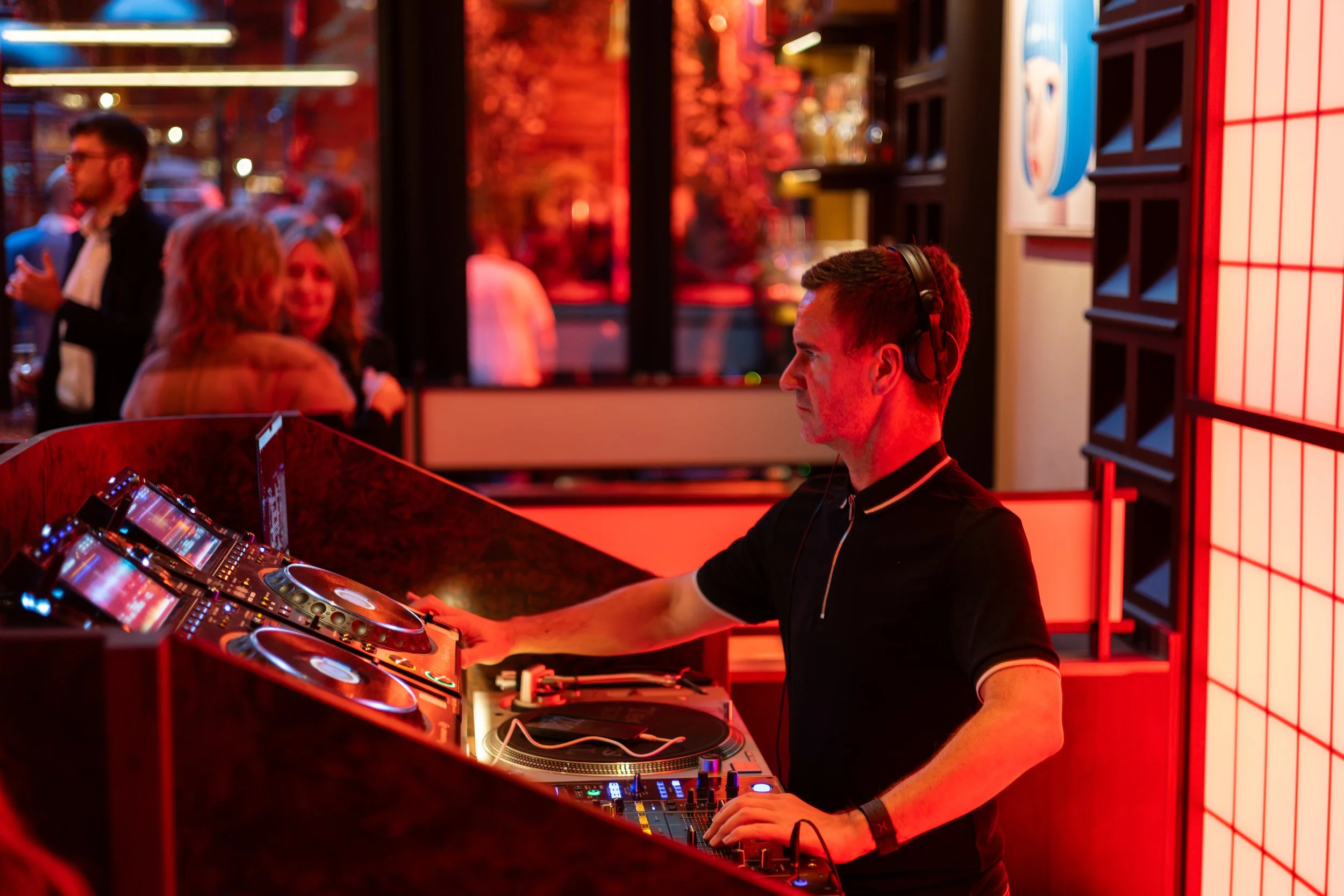 A DJ wearing headphones and a black shirt playing music at a nightclub with red lighting, while people in the background socialize and dance.