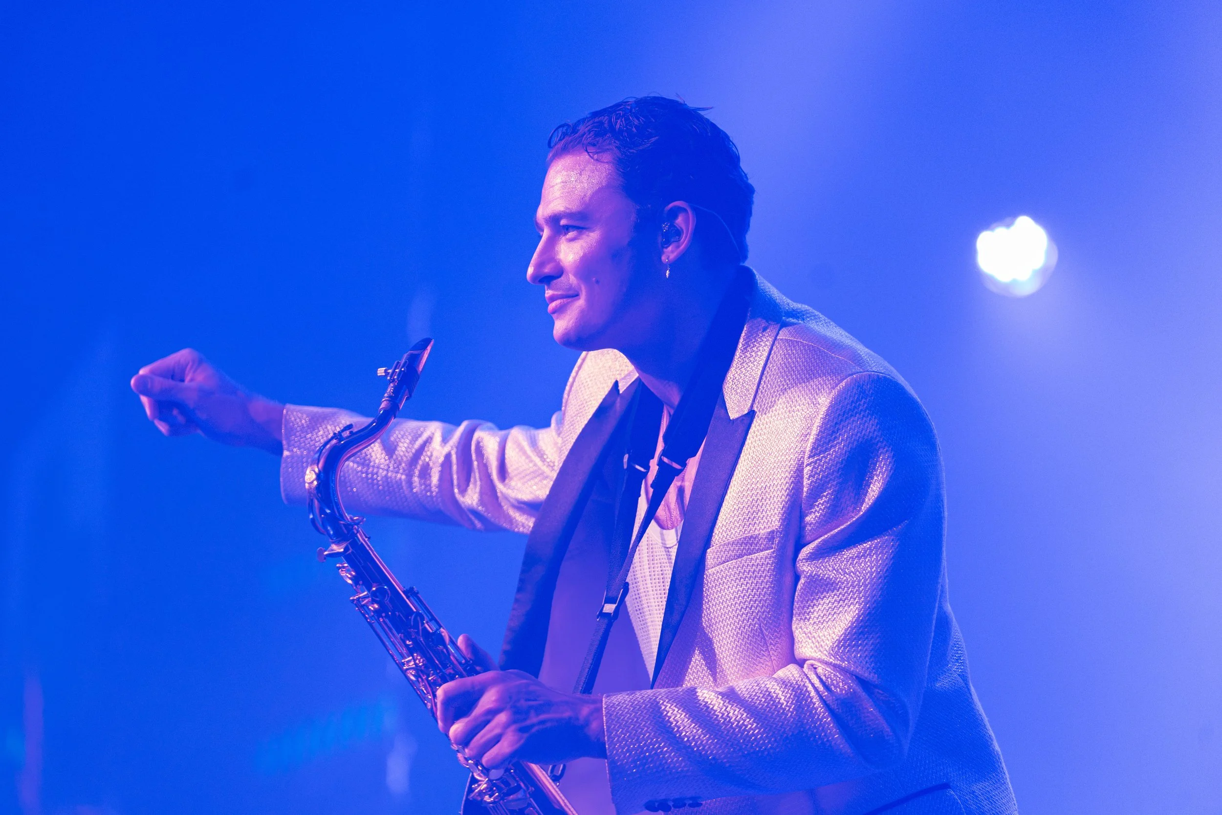 Side view of a man in a shiny gold jacket playing a saxophone on stage with blue lighting.