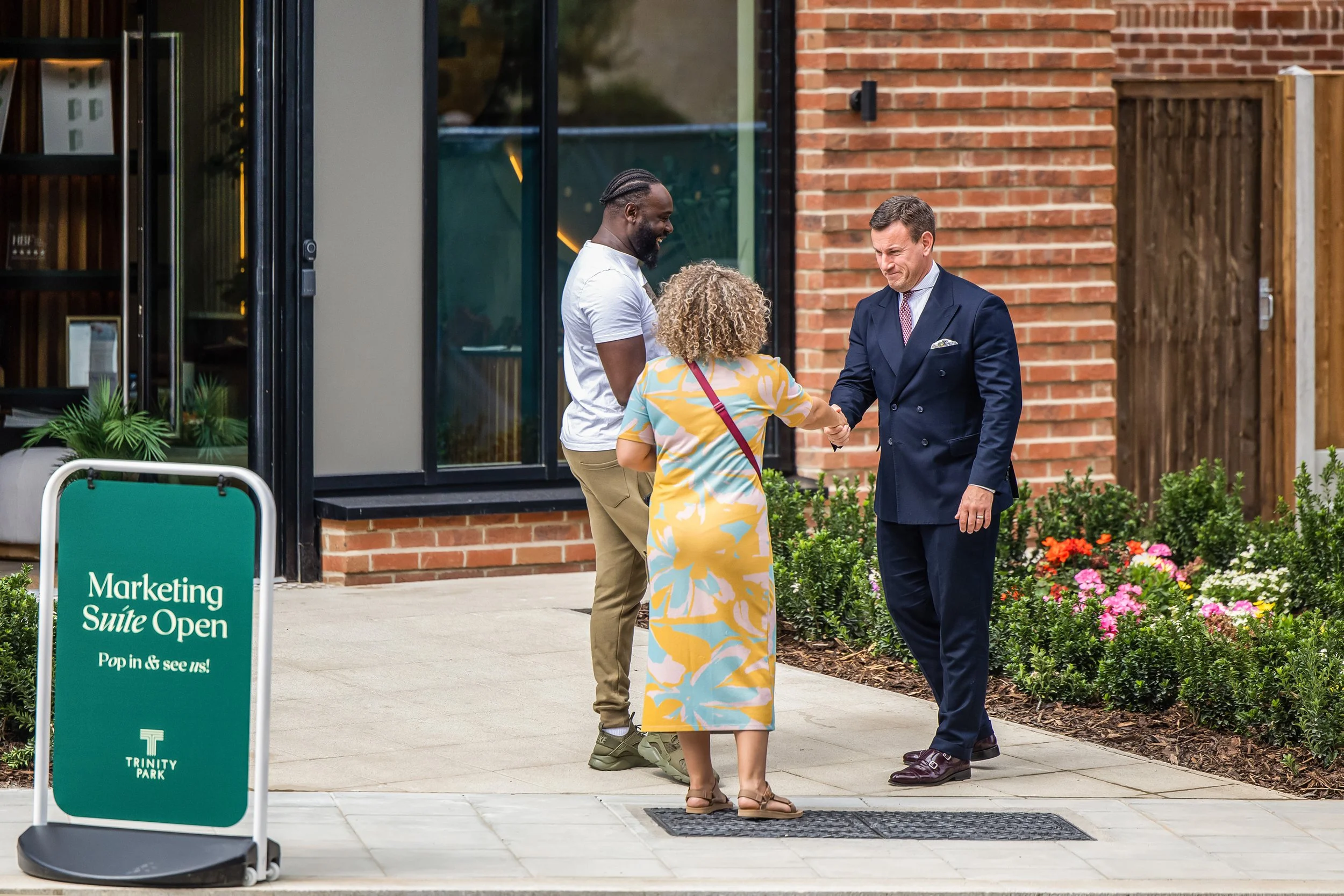 A man in a dark blue suit shaking hands with a woman in a colorful dress while another man in a white t-shirt watches. They are standing outside a building with brick walls and glass windows, near flowers and a green sign that reads 'Marketing Suite Open'.