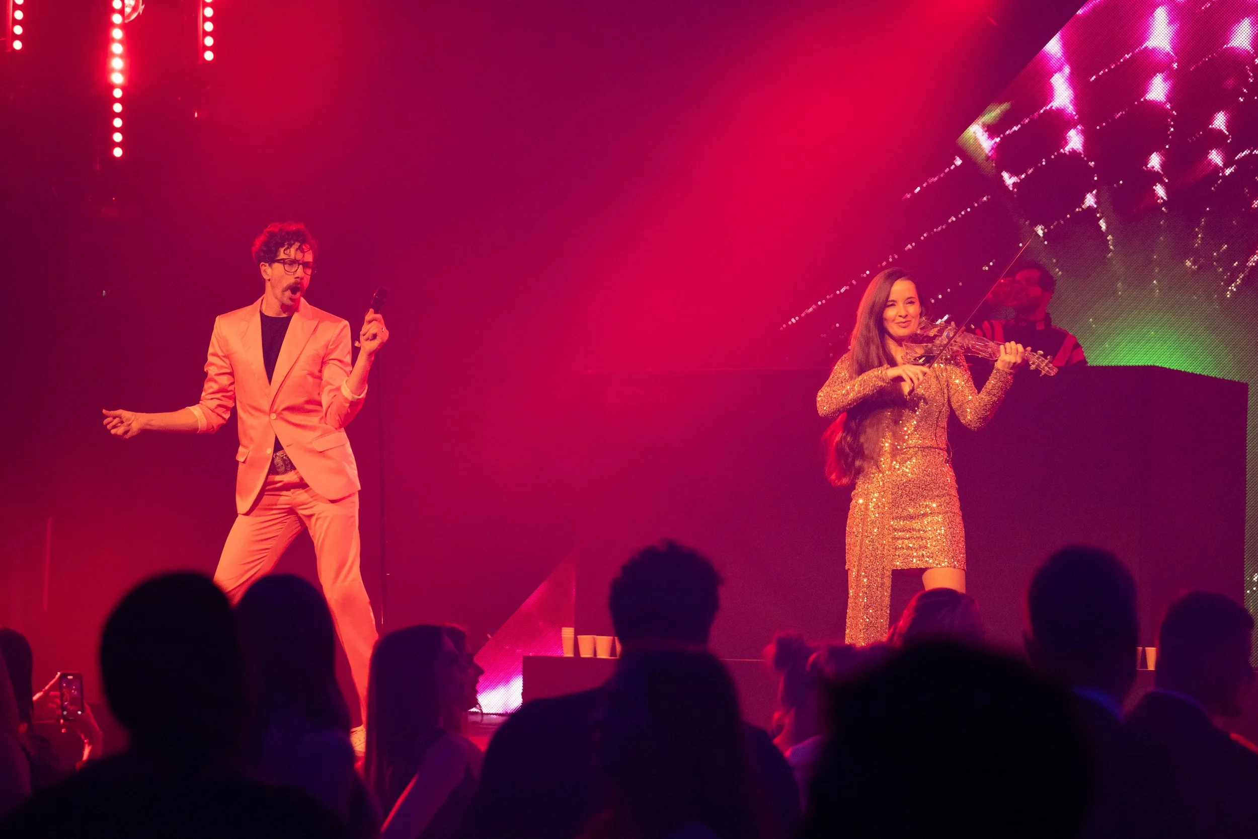 A male performer in a pink suit singing into a microphone on stage, and a female violinist in a shiny gold dress playing her instrument, with an audience in front enjoying the performance.
