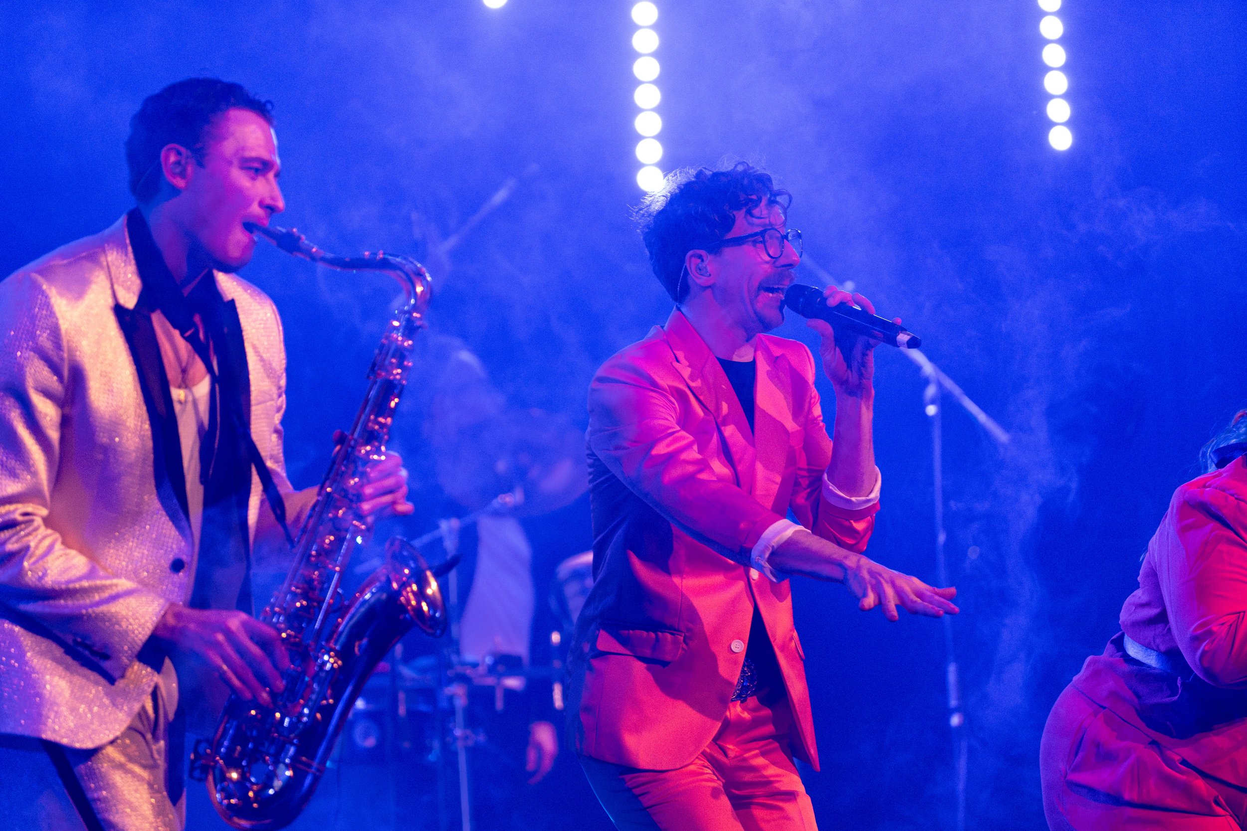 A male saxophonist in a shiny gold jacket playing a saxophone, and a male singer in a pink blazer singing into a microphone, performing on stage with colorful lights.