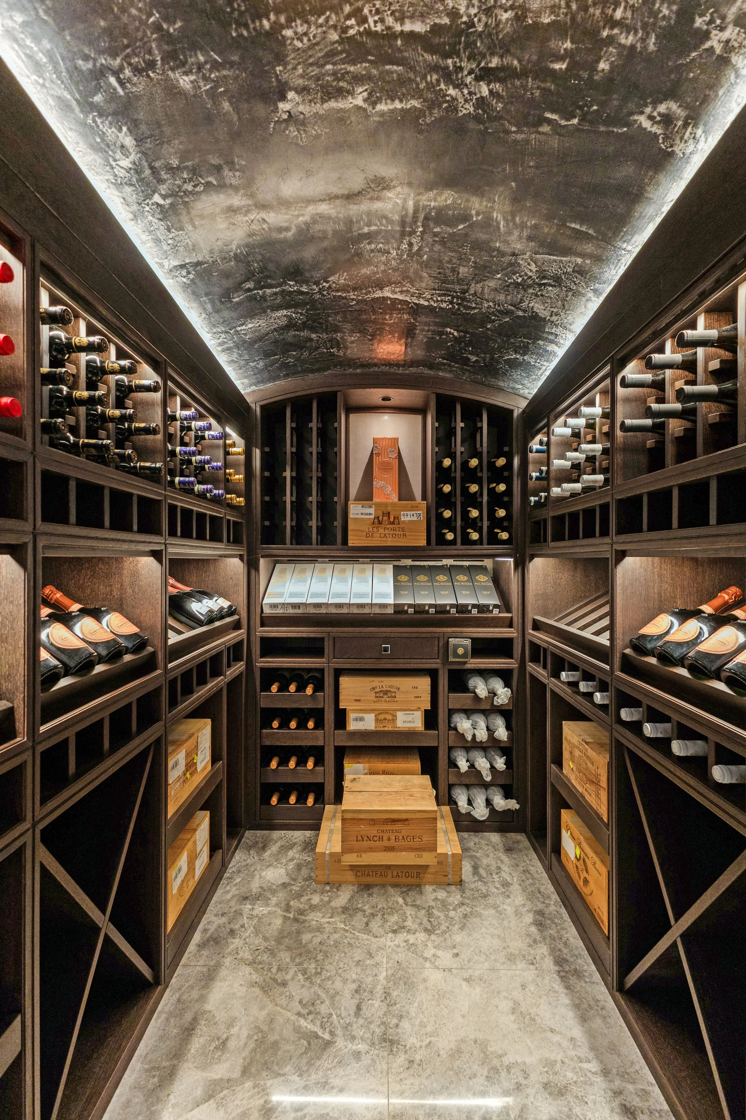 A wine cellar with dark wooden shelving on both sides and a gray stone floor. Bottles of wine are stored on the shelves, some in wooden boxes. The ceiling has a textured, gray finish with lighting around the edges. At the end of the aisle, there is a wooden box and some wine bottles stored on the lower shelves.