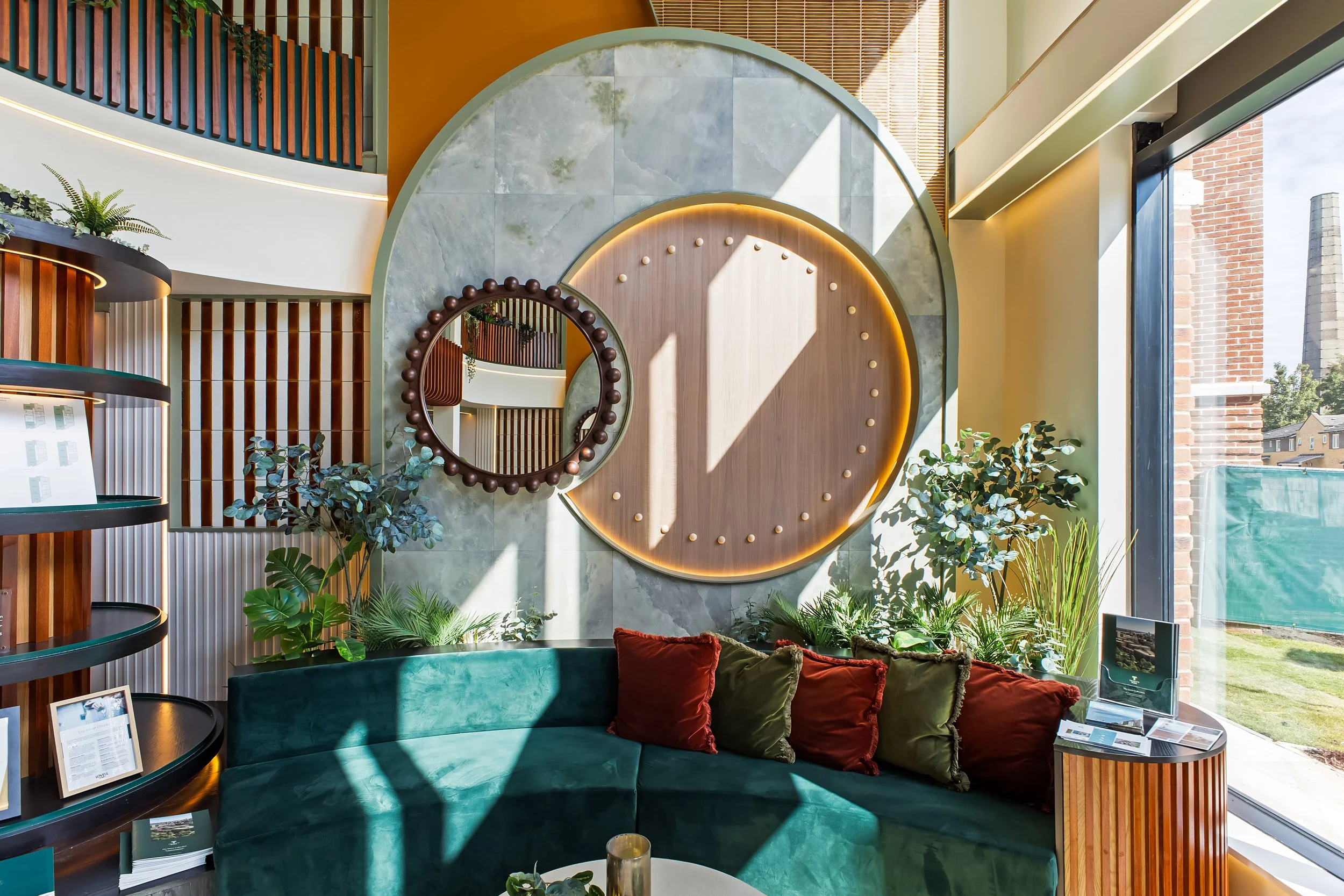 Interior of a modern lounge or waiting area with a curved green velvet sofa with red and dark green throw pillows, a small round coffee table, and a bookshelf with plants and framed photos. The wall features a large circular mirror with decorative wooden accents and a backdrop of green marble tiles, with sunlight streaming through a large window showing a cityscape and a brick building outside.