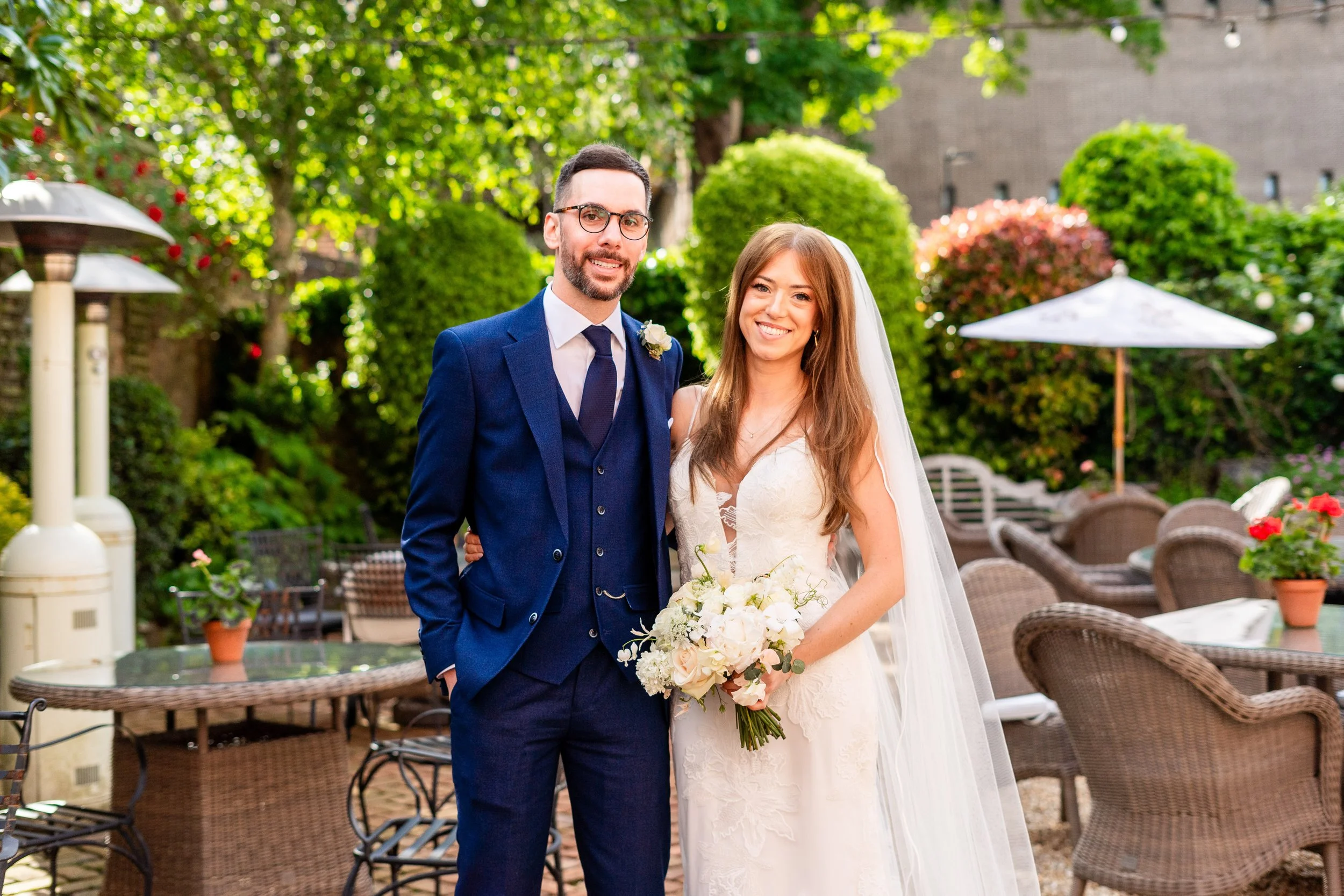 A bride and groom standing outdoors in a garden setting during daytime, smiling for a photo. The groom is wearing a blue suit with a white shirt and dark tie, and the bride is dressed in a white wedding gown holding a bouquet of white flowers. The background features green trees, colorful bushes, patio furniture, and large white patio umbrellas.