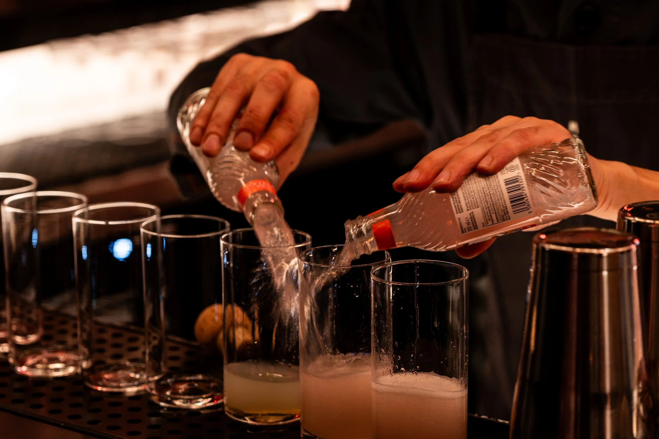 Hand pouring clear liquid from a bottle into a row of empty shot glasses on a bar counter, with a dark background and warm lighting.