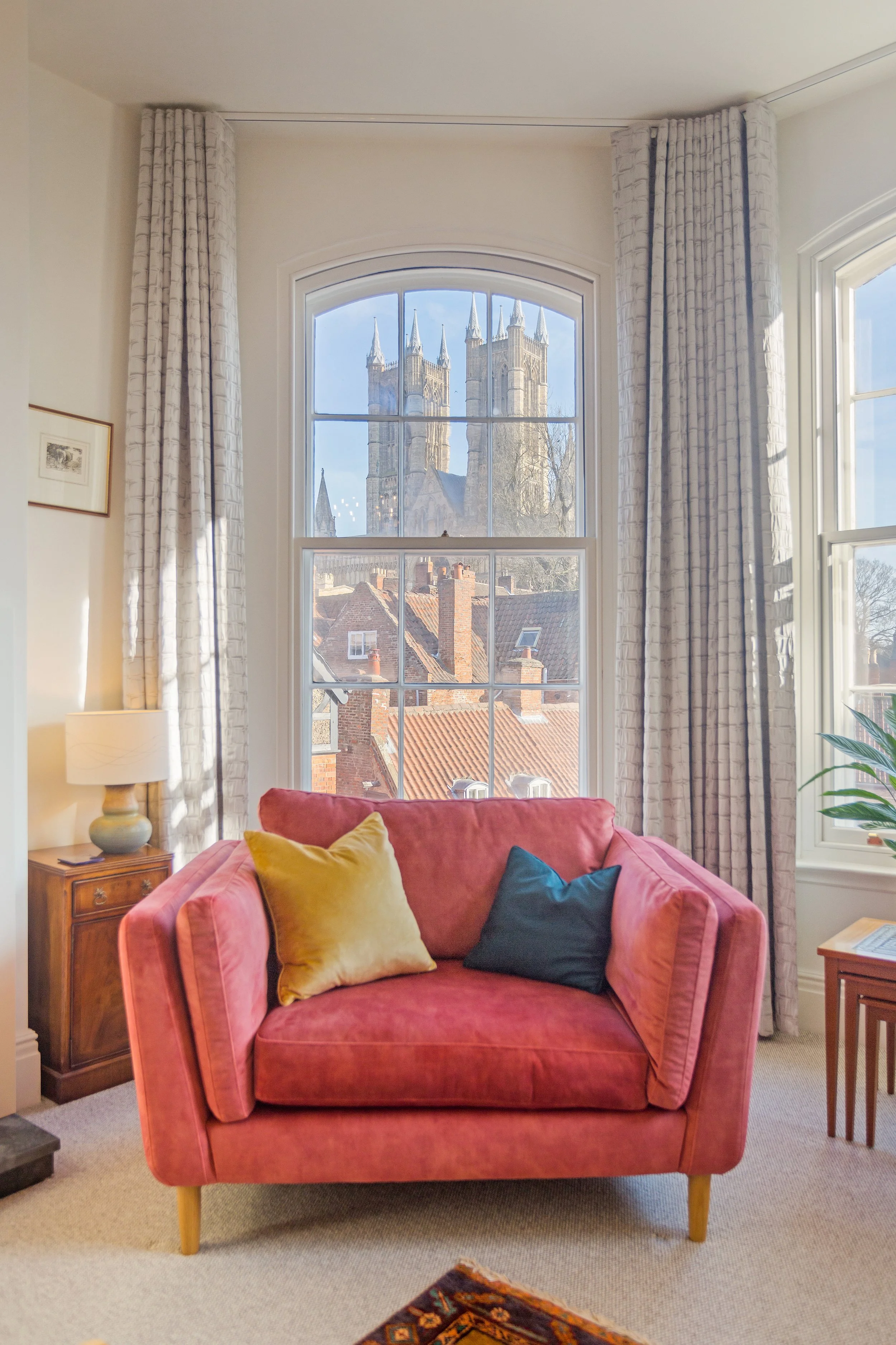 A cozy sitting room with a salmon-colored sofa, embroidered cushions, a wooden side table with a lamp, and large windows revealing historic Romanesque-style churches with tall spires outside.