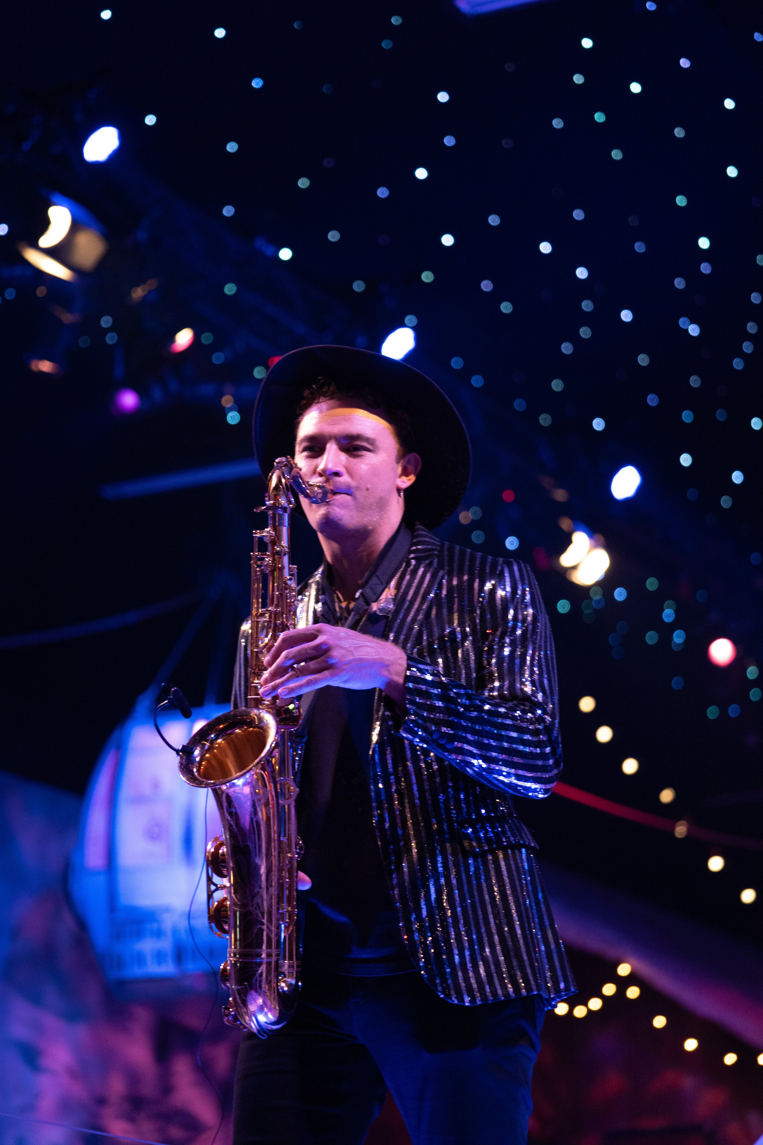 A man playing the saxophone on a stage with colorful, out-of-focus lights in the background.