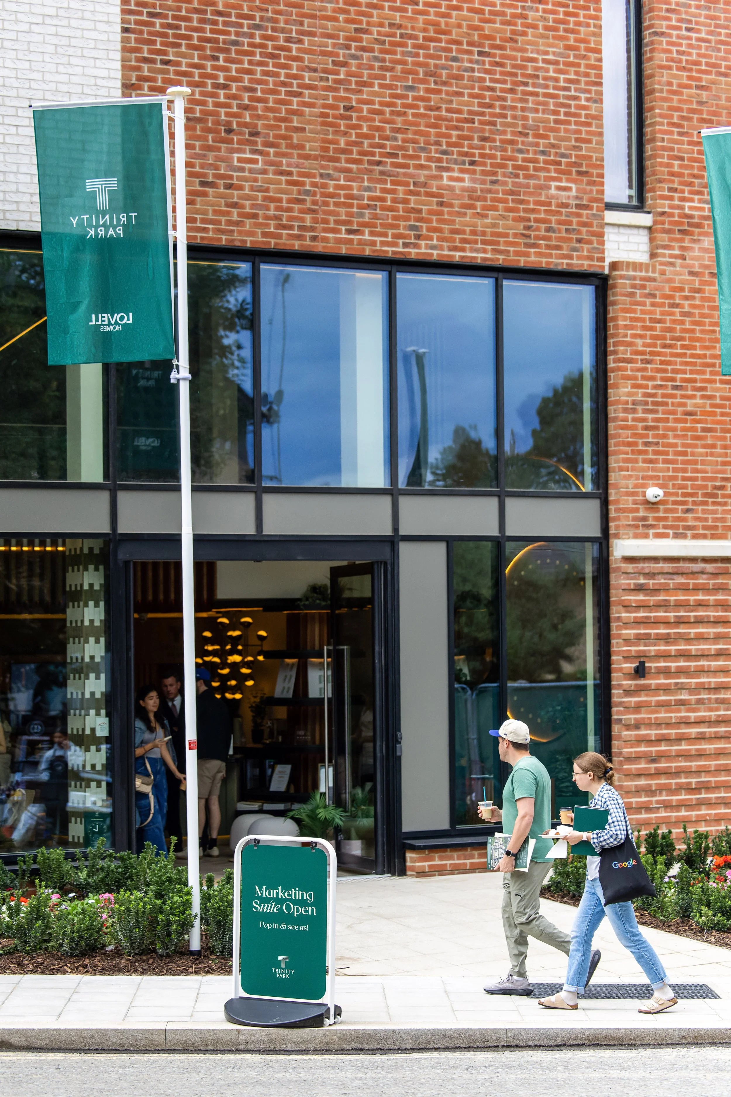 People walking past a modern brick building with large glass windows, a green sign on a stand advertising a marketing suite open, and visitors inside the building.