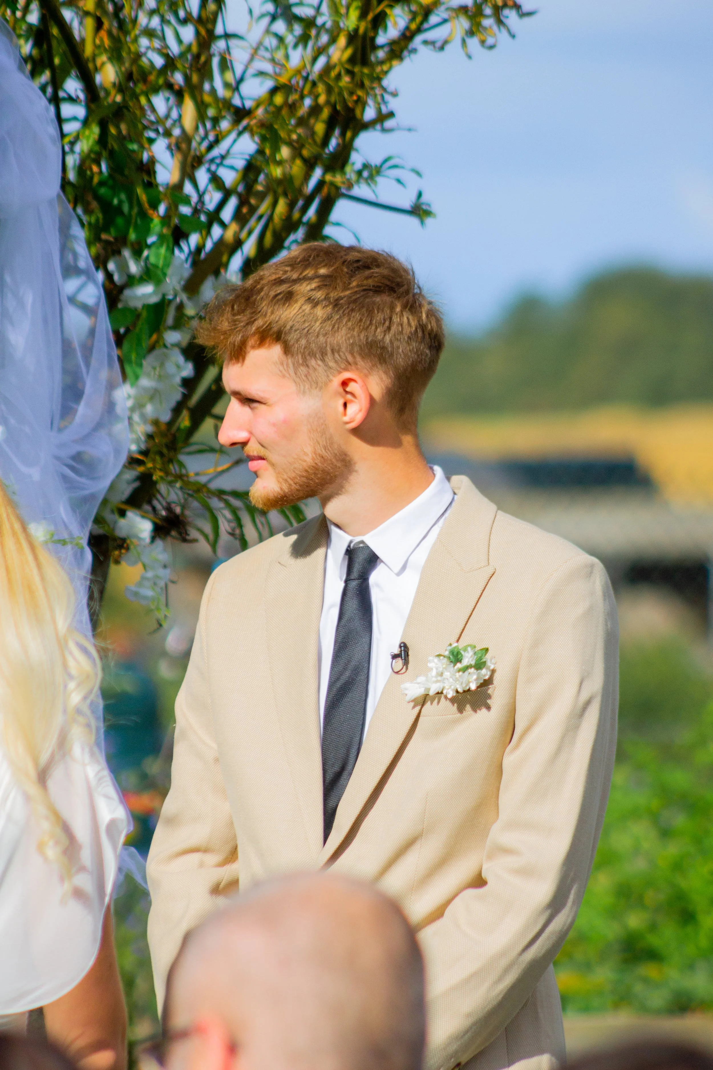 A man with light brown hair and a beard dressed in a beige suit and white shirt, standing outdoors near greenery, looking to the left.