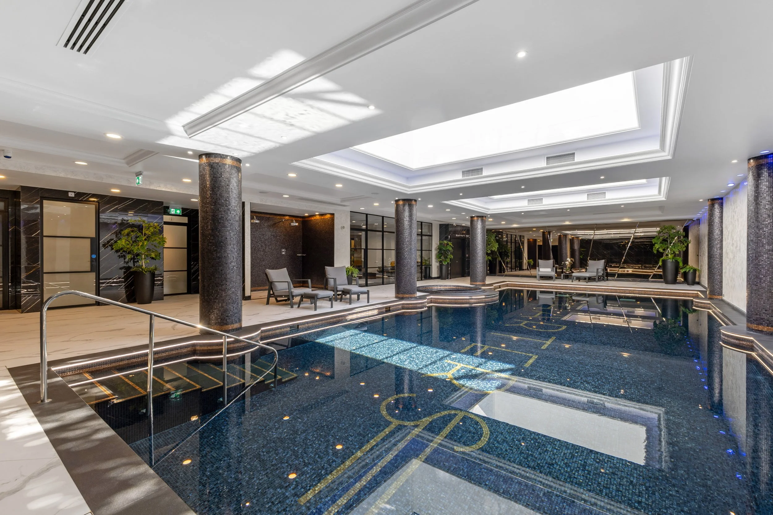 Luxurious indoor pool area with black tiled pool, white marble flooring, black mosaic columns, and modern seating with potted plants, illuminated by ceiling lights.