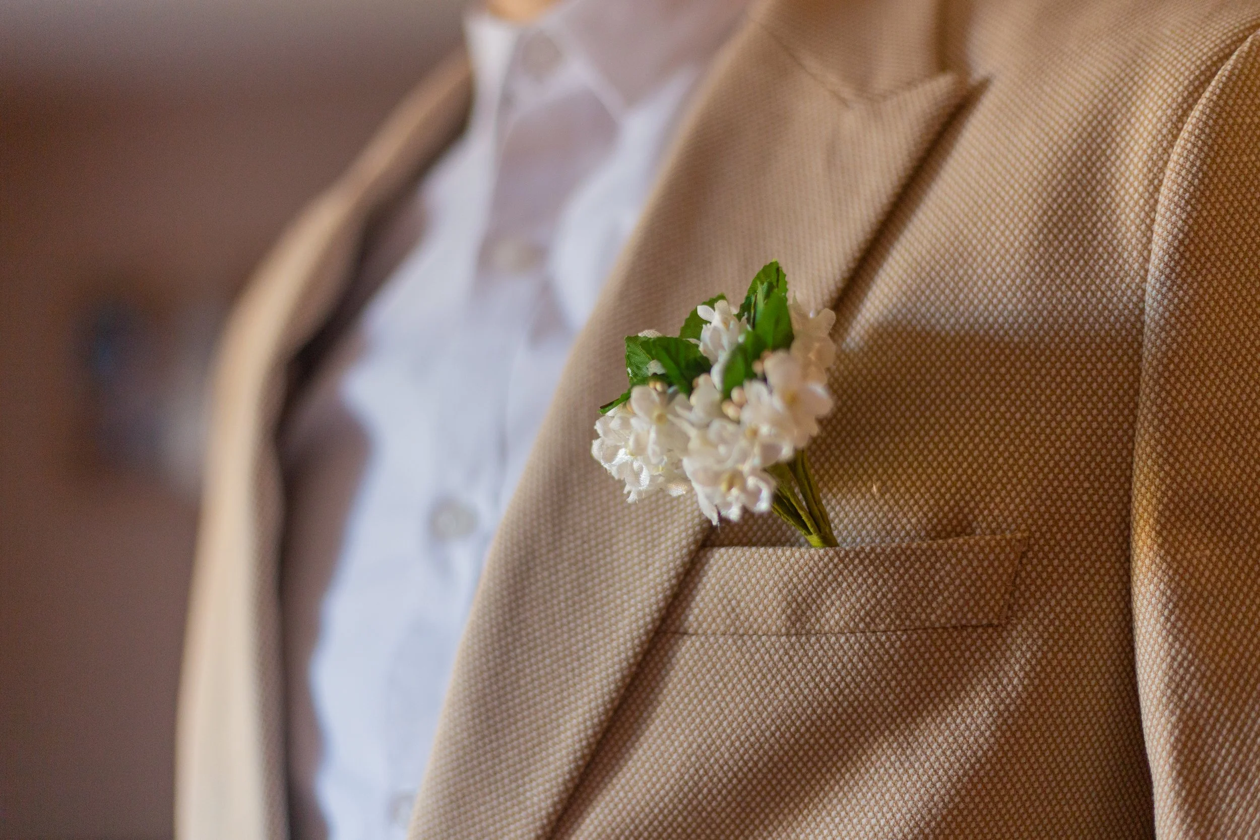 Close-up of a man wearing a beige suit with a white shirt and a boutonniere of small white flowers with green leaves on the lapel.