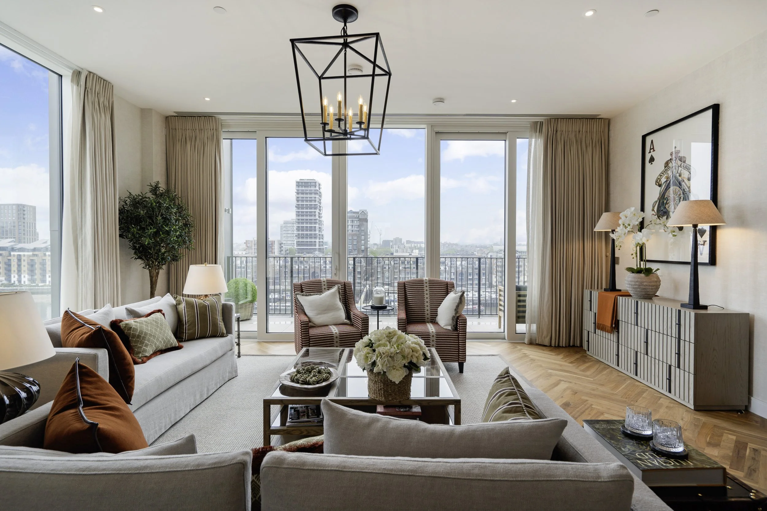 Living room with large windows, beige curtains, a chandelier, a white sofa with pillows, armchairs with striped fabric, a coffee table with flowers, and wall art of a playing card.