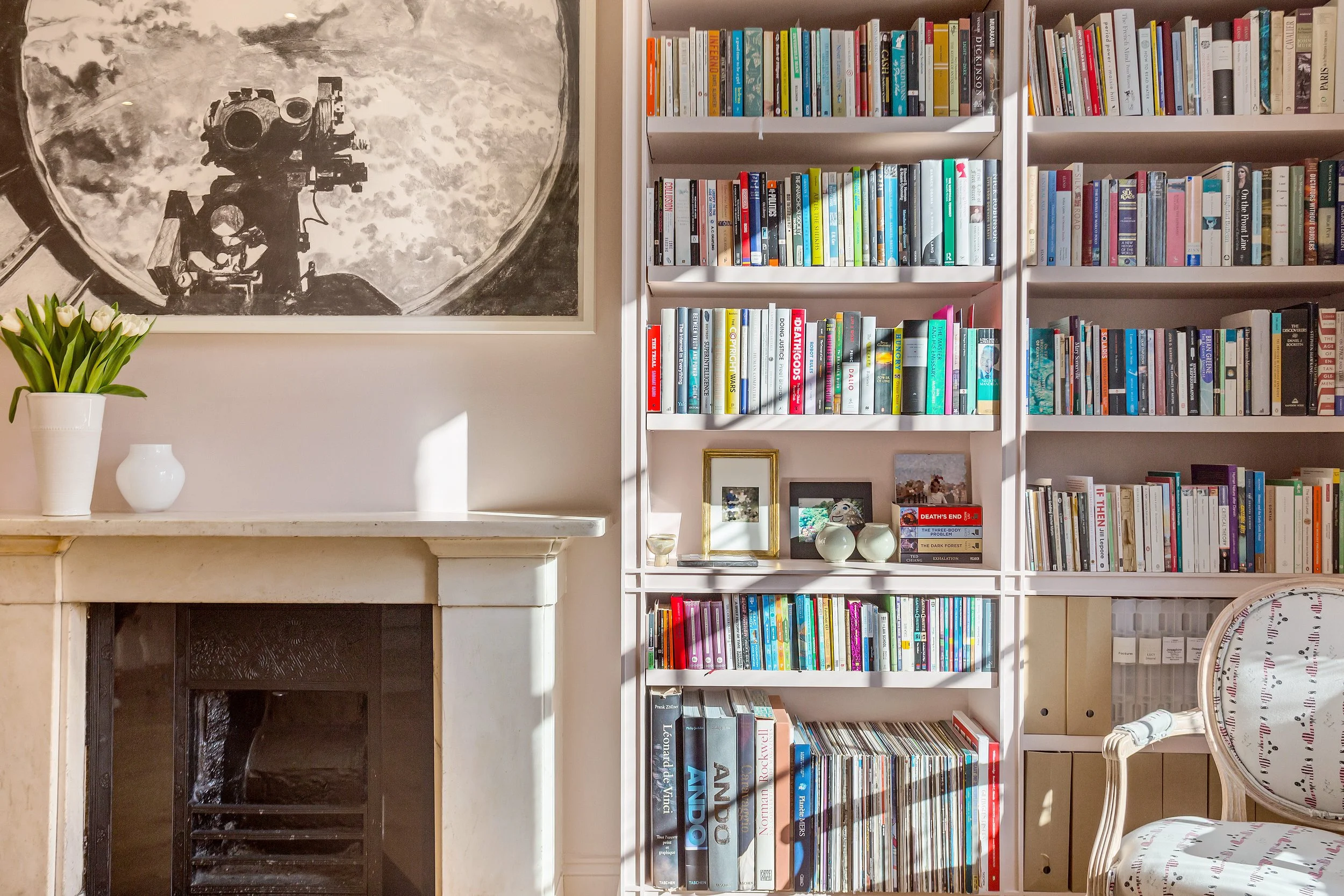 Living room with a fireplace on the left and white bookshelves filled with books on the right, a framed picture and small decorative objects on the shelves, and a chair with a floral pattern on the bottom right.
