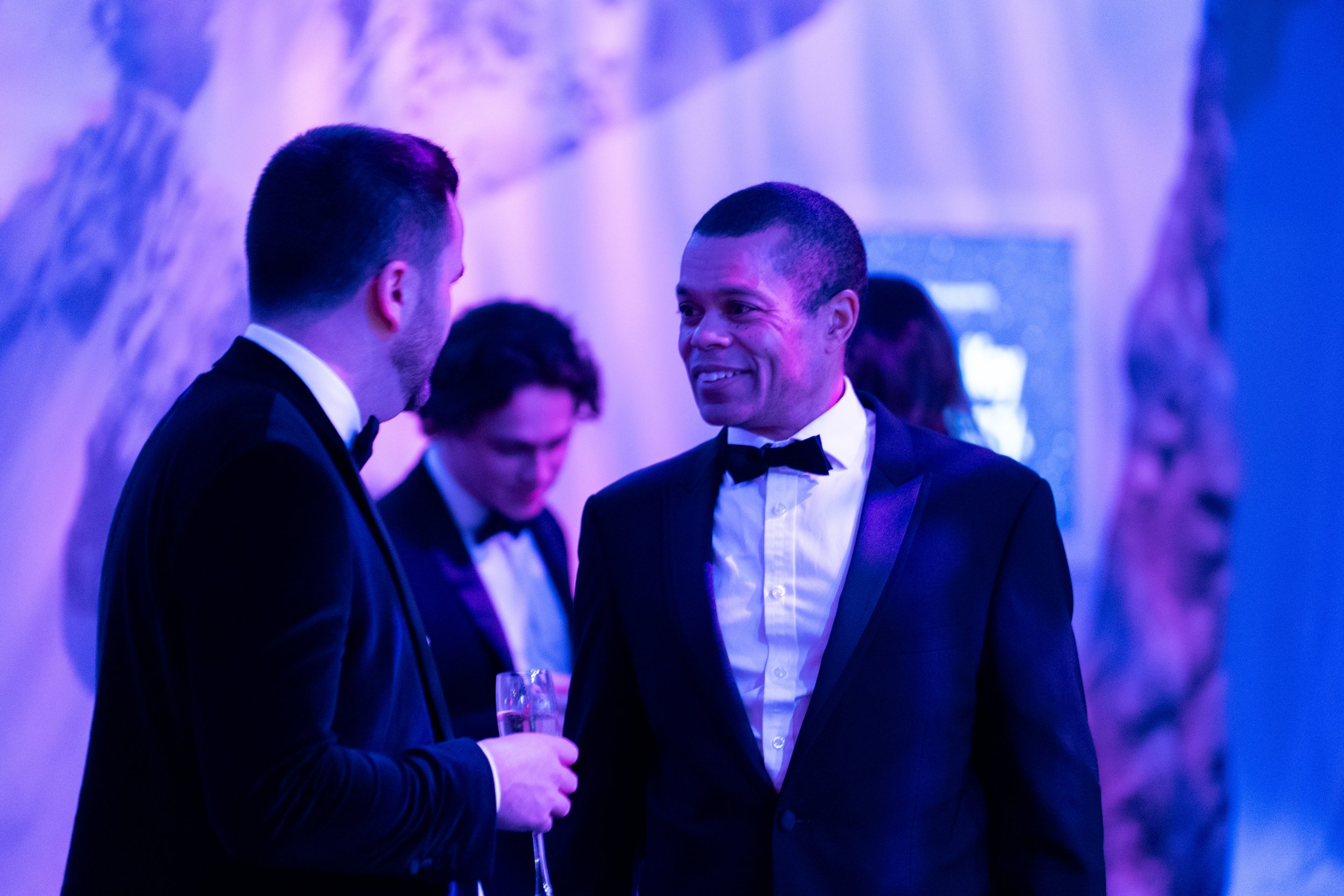 Two men in tuxedos talking at a formal event with purple lighting. One man holds a champagne flute, smiling, while the other appears to be engaged in conversation. In the background, there are other guests also dressed formally.