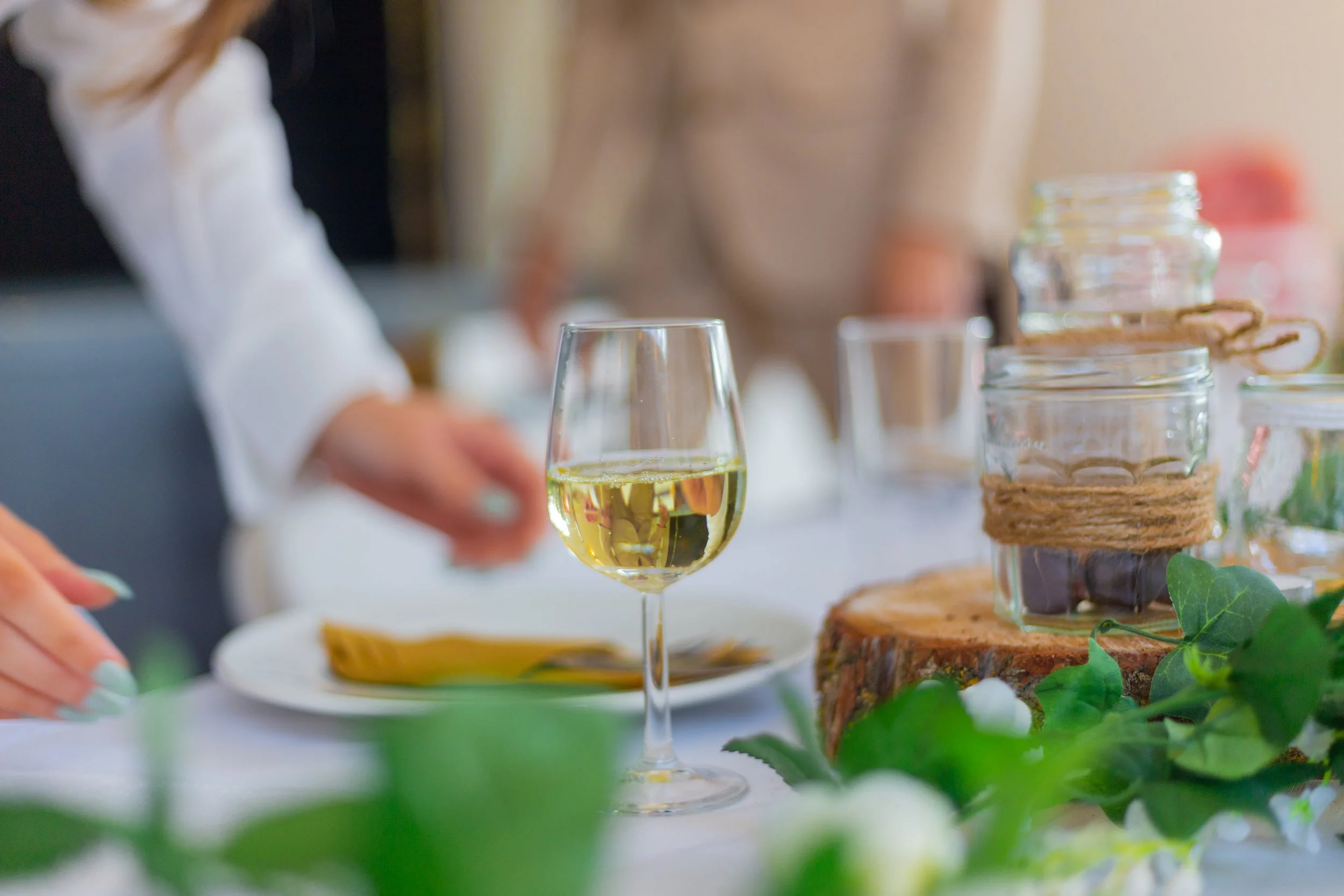 A glass of white wine on a table with various jars and greenery in the background.