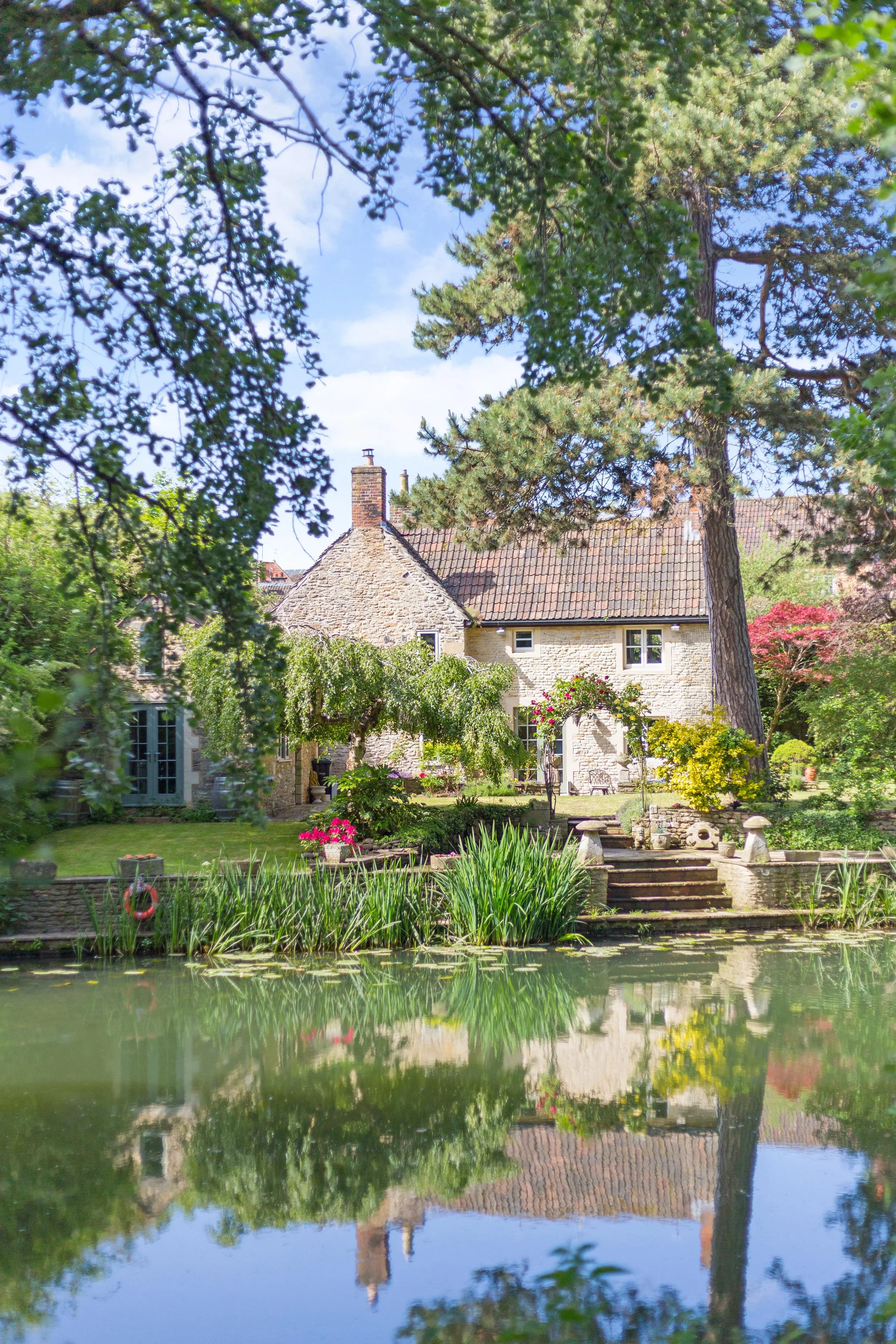 A cozy stone house with a red chimney surrounded by lush garden greenery, trees, and a pond with reflections of the house and trees under a partly cloudy sky.