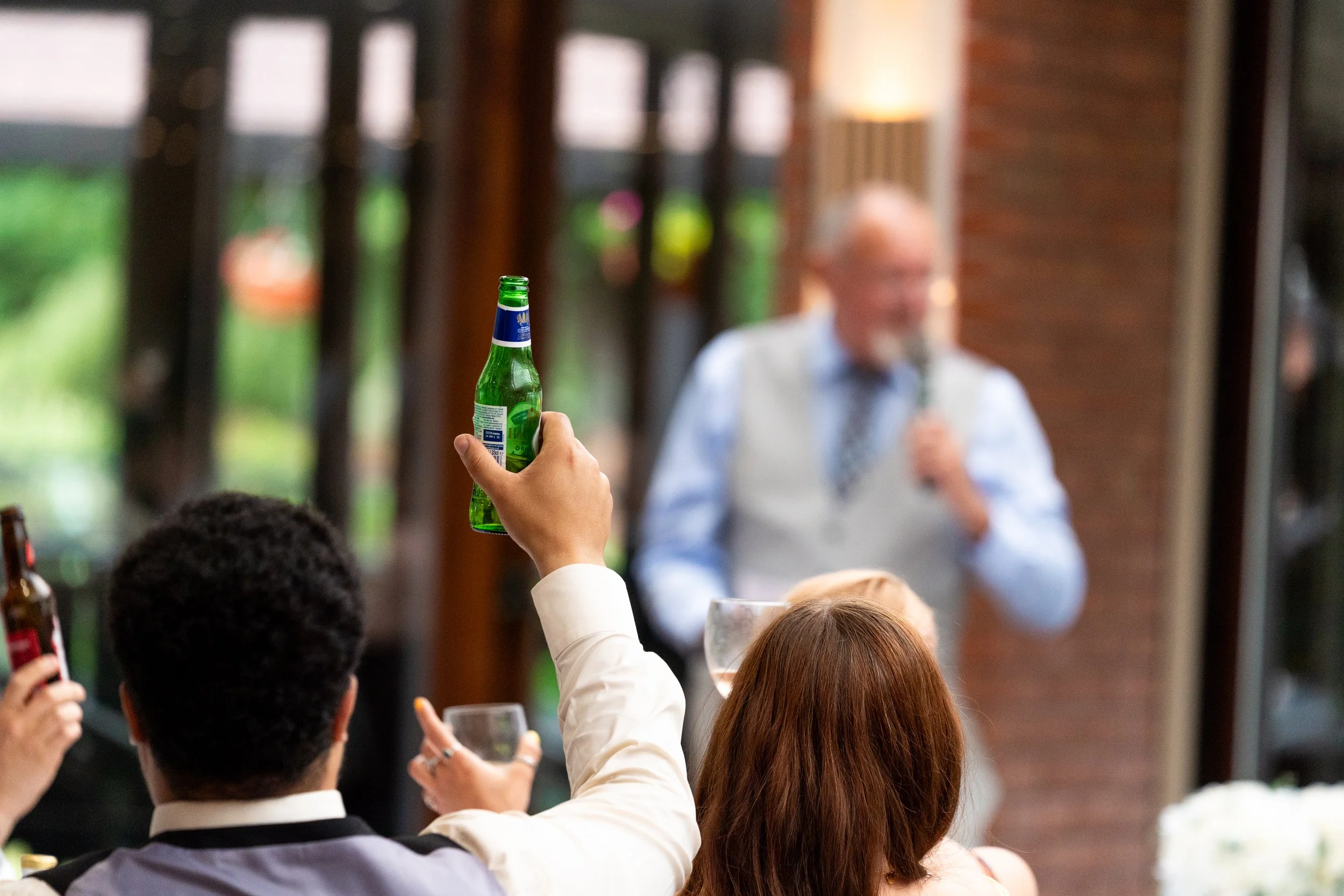 Person holding a green bottle at a social gathering with people sitting around a table, a man with glasses standing and speaking in the background.