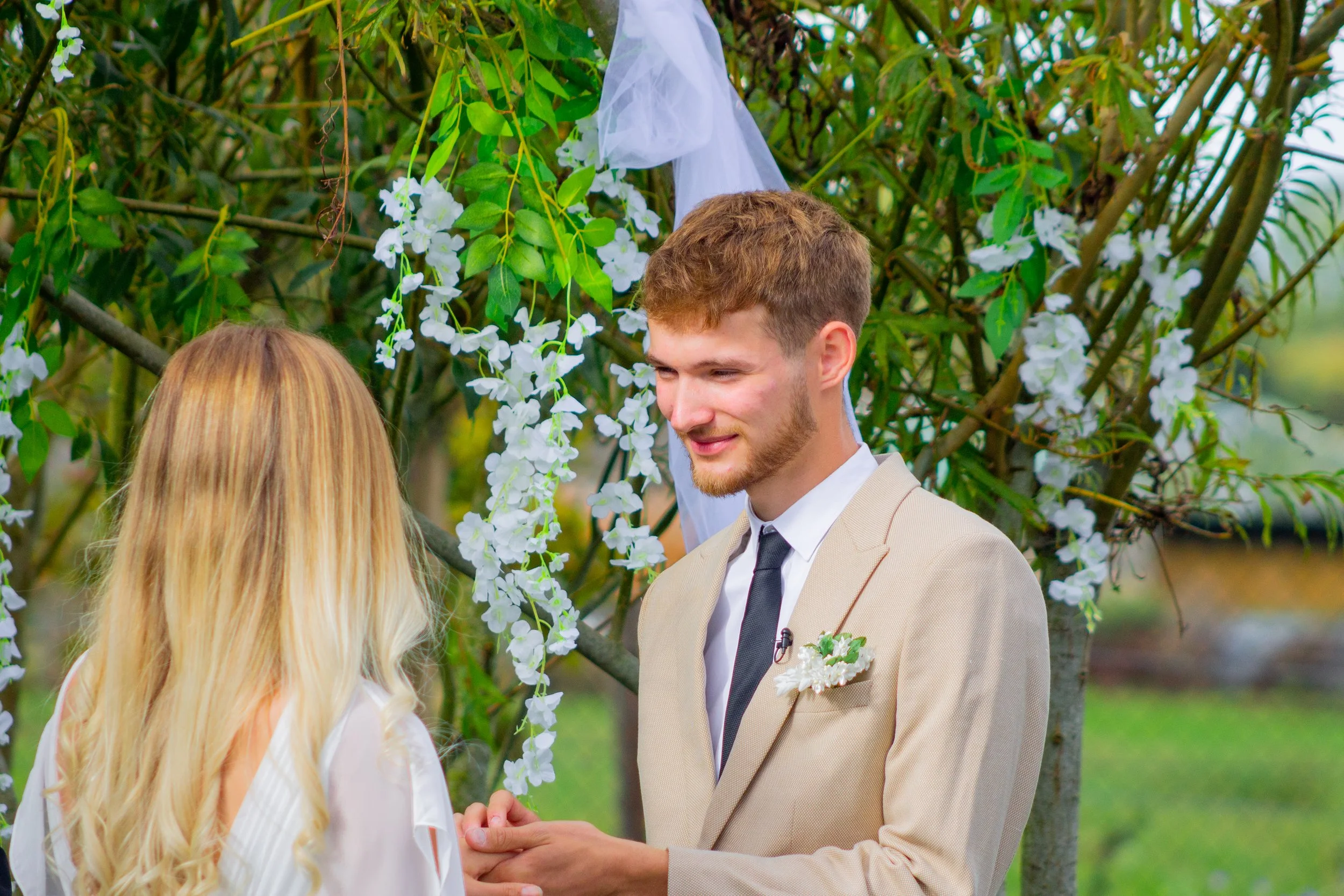 A man and woman exchanging wedding vows outdoors under a tree decorated with white flowers and fabric during a wedding ceremony.