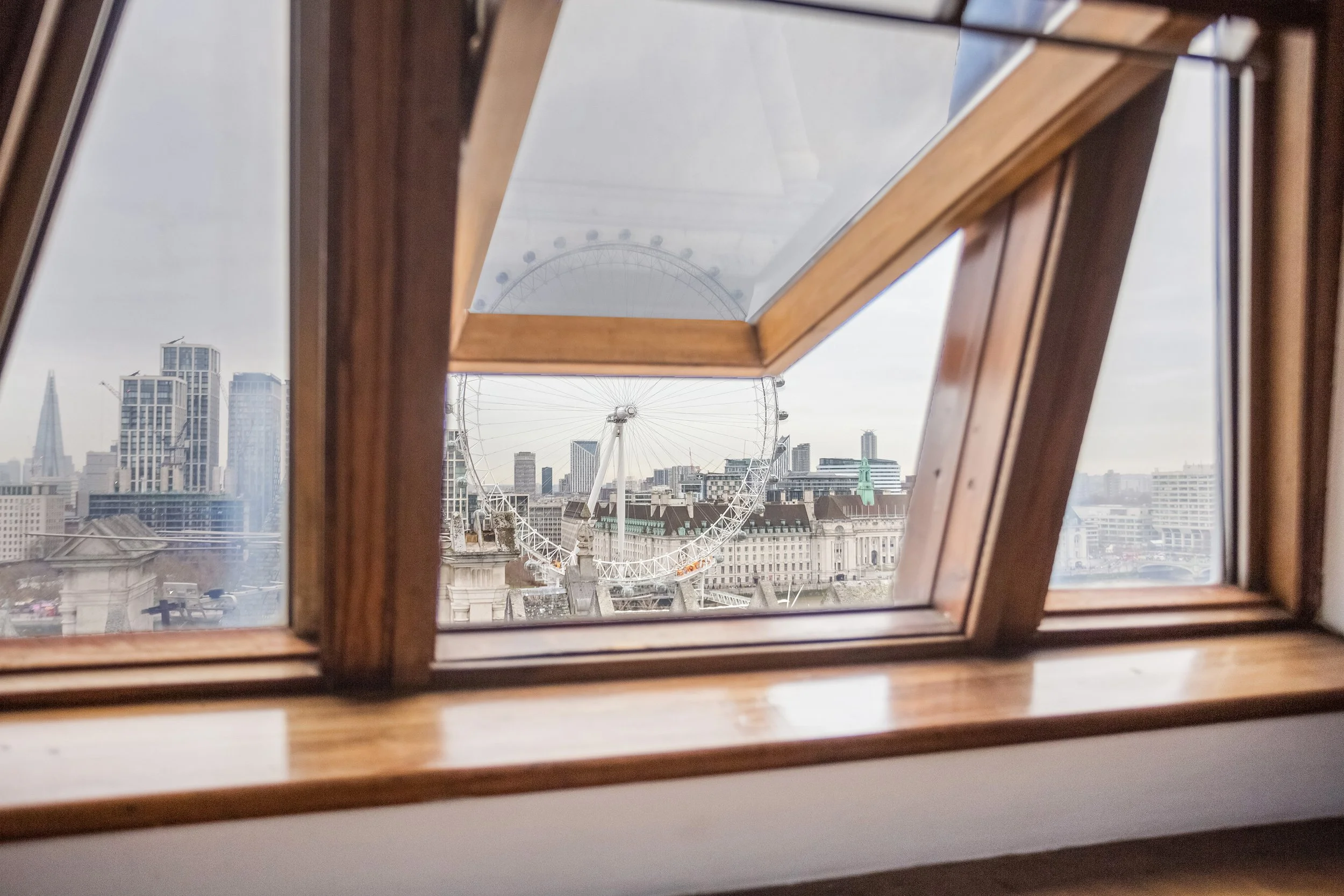 View through a window showing the London Eye Ferris wheel and a cityscape of buildings under a cloudy sky.