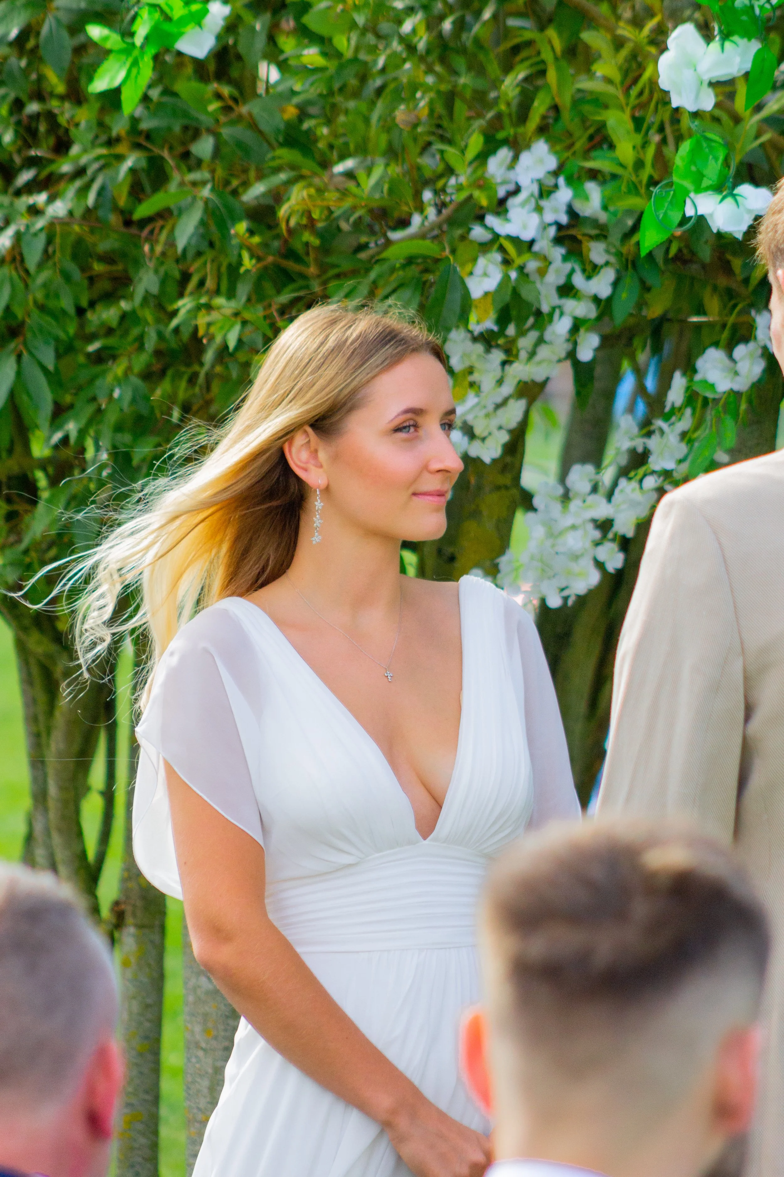 A woman with long blonde hair wearing a white dress and jewelry appears to be participating in a ceremony outdoors, with greenery and white flowers in the background. Several children are in the foreground, and an individual in a light-colored suit is partially visible on the right.