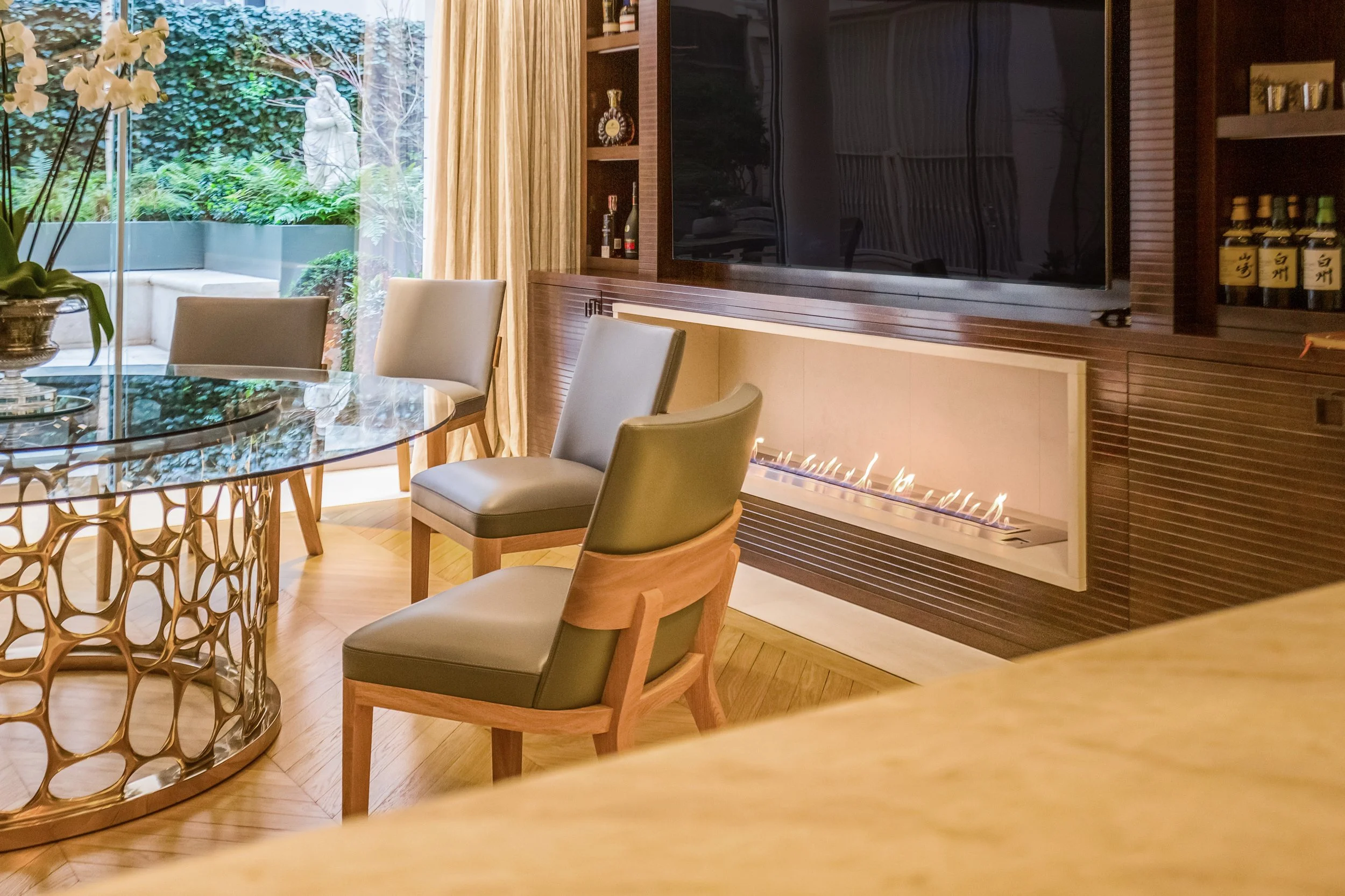 Contemporary living room with a glass-topped dining table, beige upholstered chairs, a wooden entertainment center with a large flat-screen TV, a modern fireplace with visible flames, and large windows with beige curtains overlooking a lush garden with trees and statues.