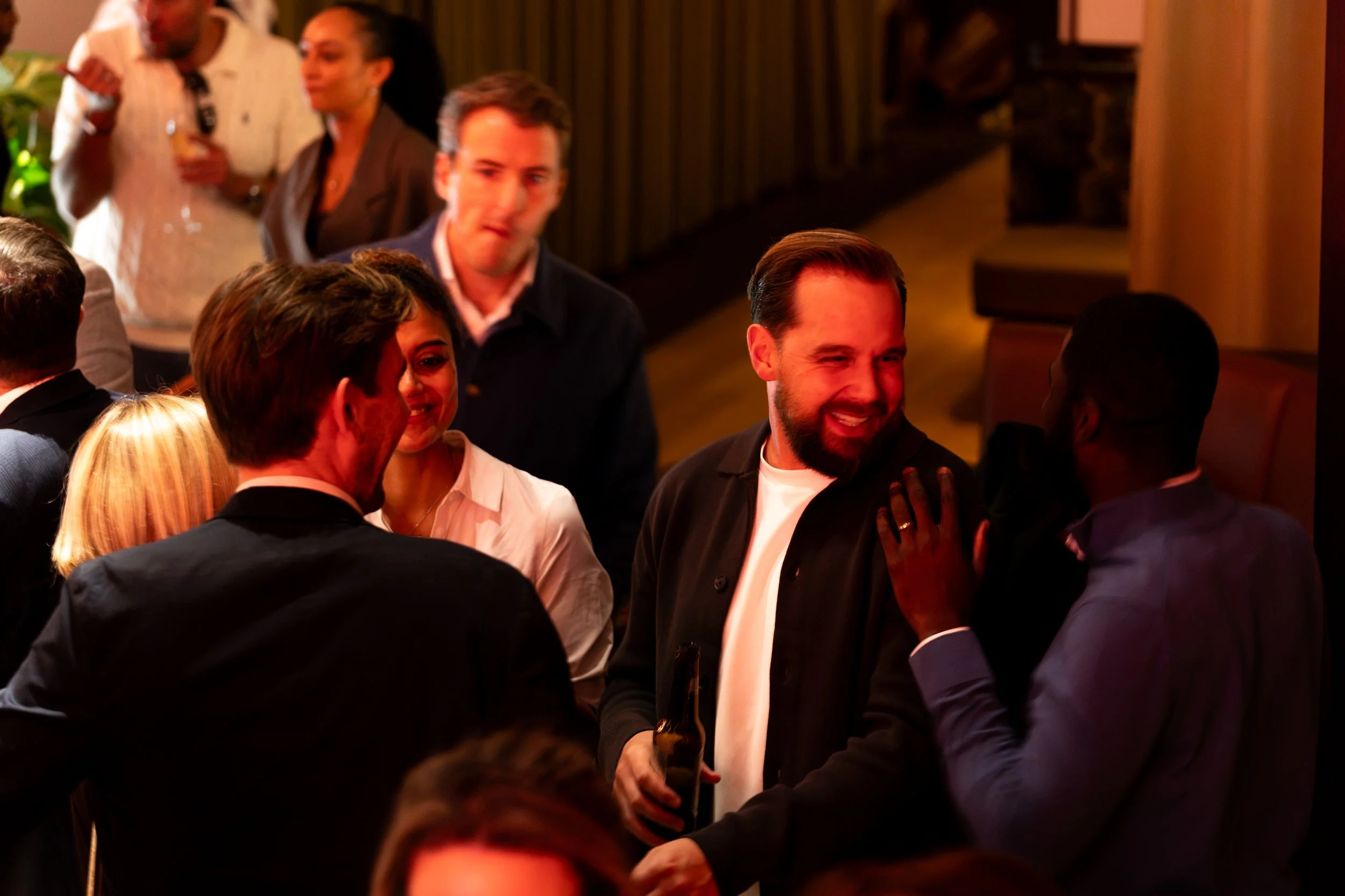 Group of people at a social gathering, smiling and talking, in a warmly lit indoor setting.