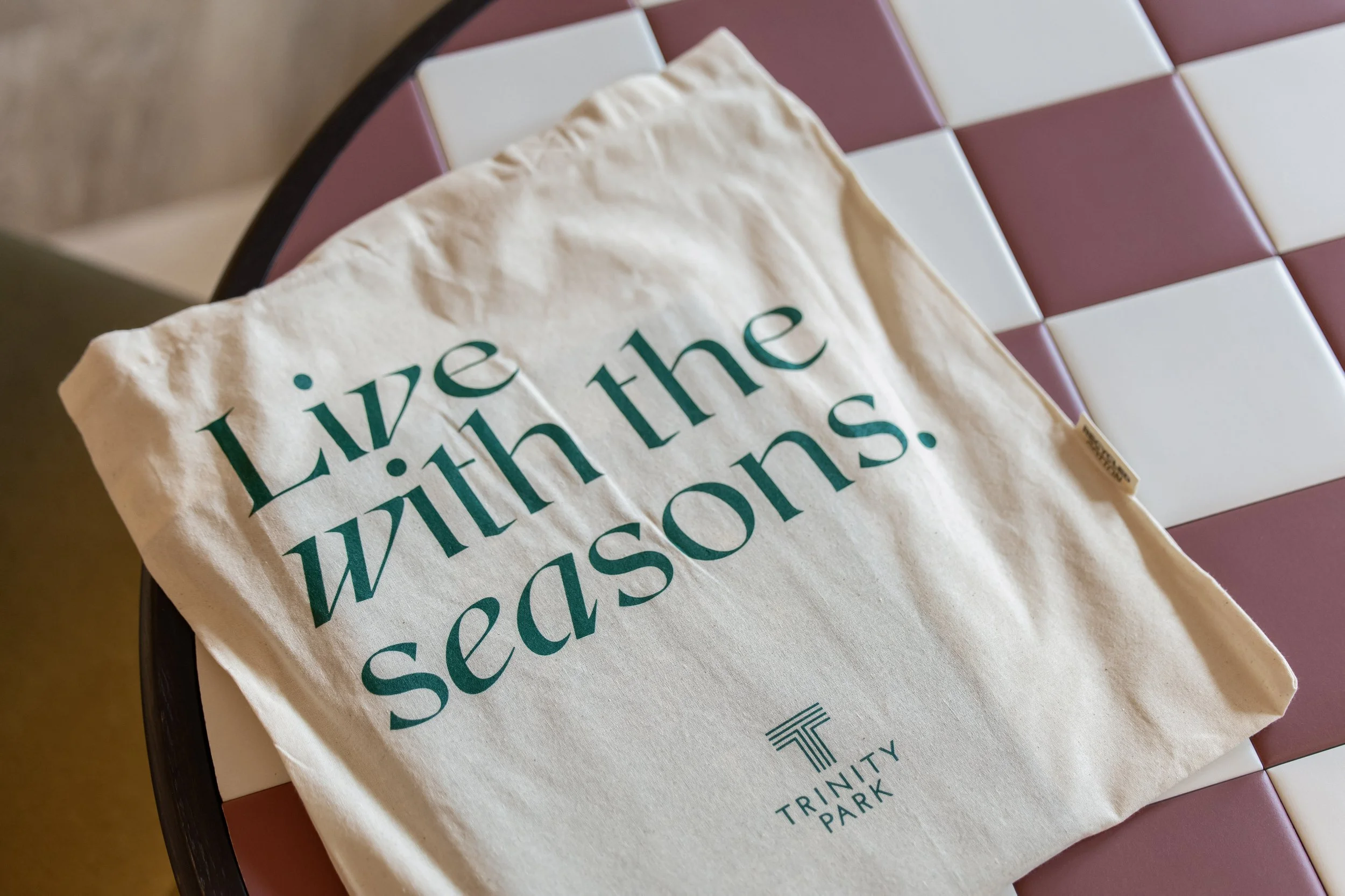 A white cloth bag with green text that reads 'Live with the seasons.' It is placed on a checkered table with burgundy and white tiles.