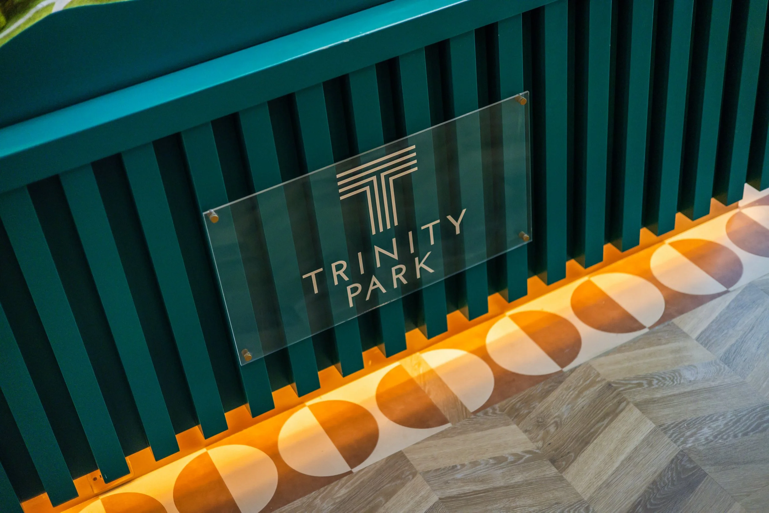 A teal-colored wooden slat wall with a clear acrylic sign that reads 'Trinity Park' and a stylized 'T' logo, illuminated by warm orange lighting at the bottom.