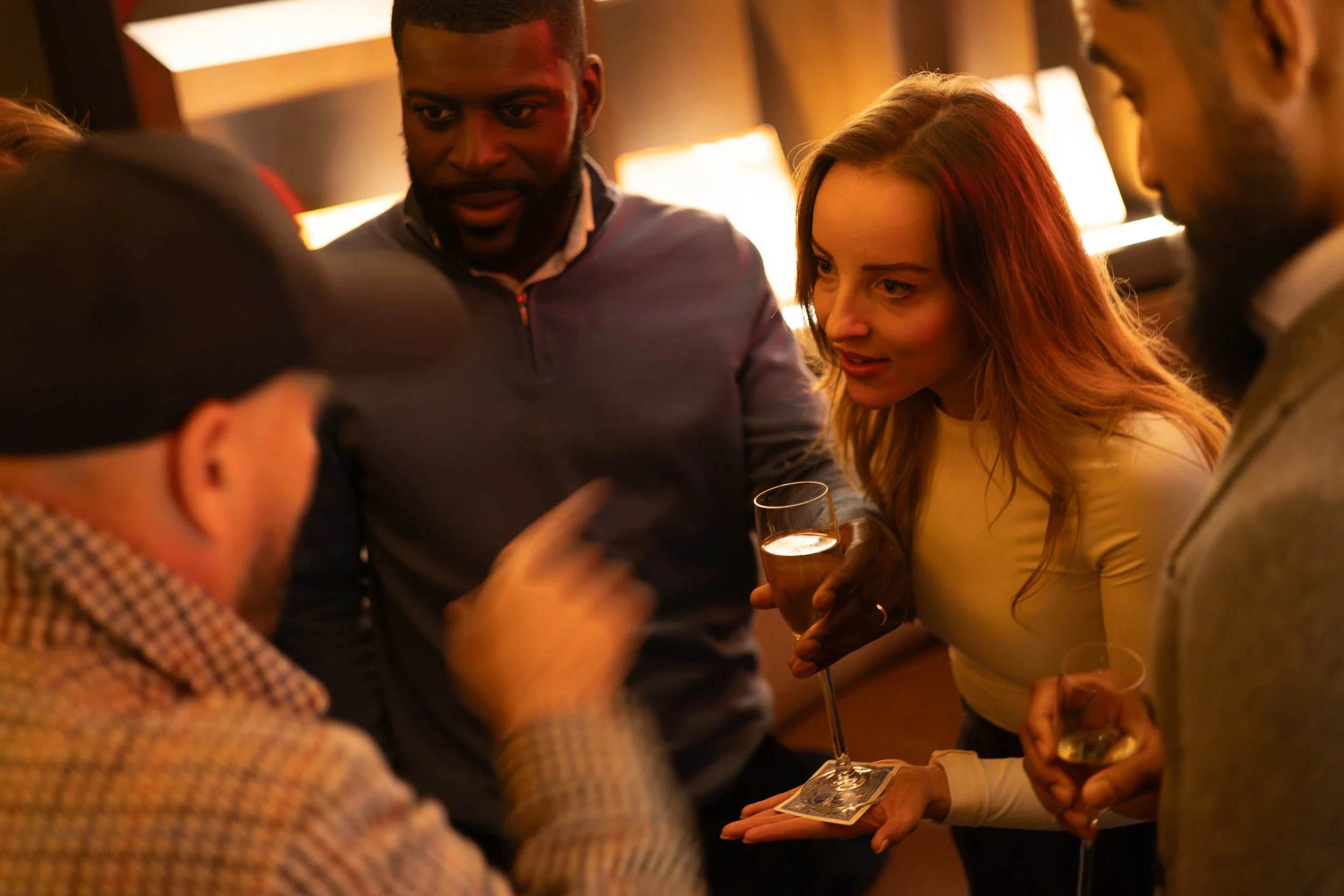 Group of people engaging in conversation at a social gathering, with some holding drinks, in a warmly lit indoor setting.