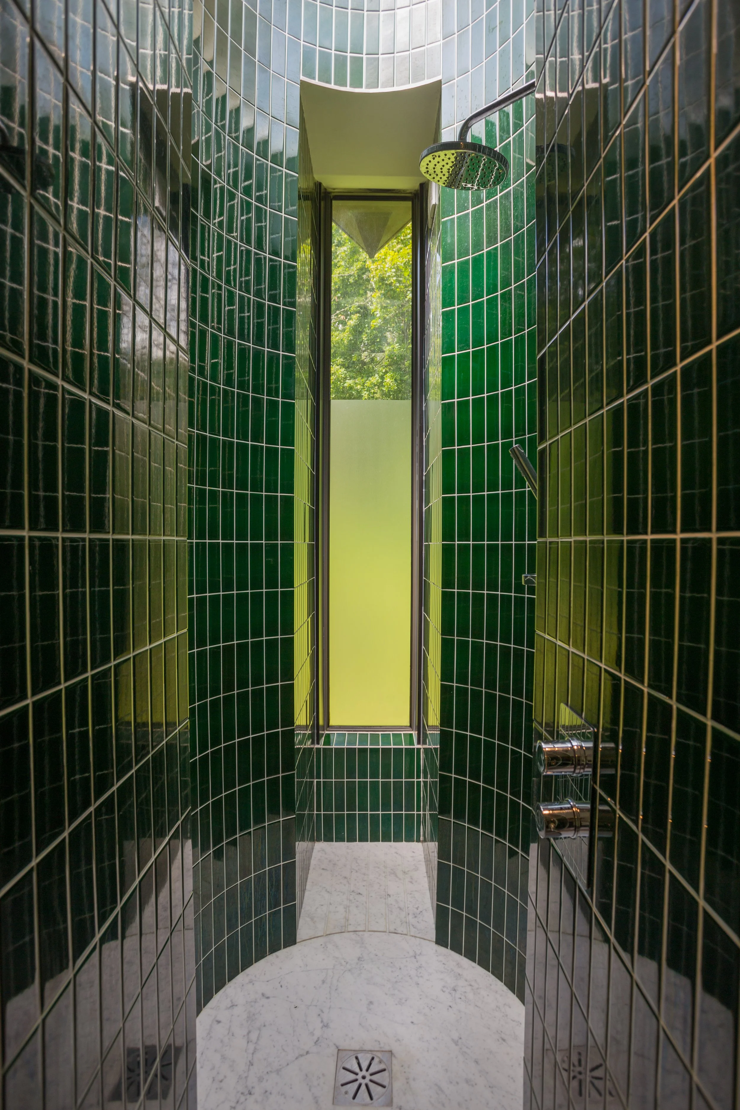 A modern shower with dark green tiled walls, a rainfall showerhead, and a glass door leading outside to green trees.