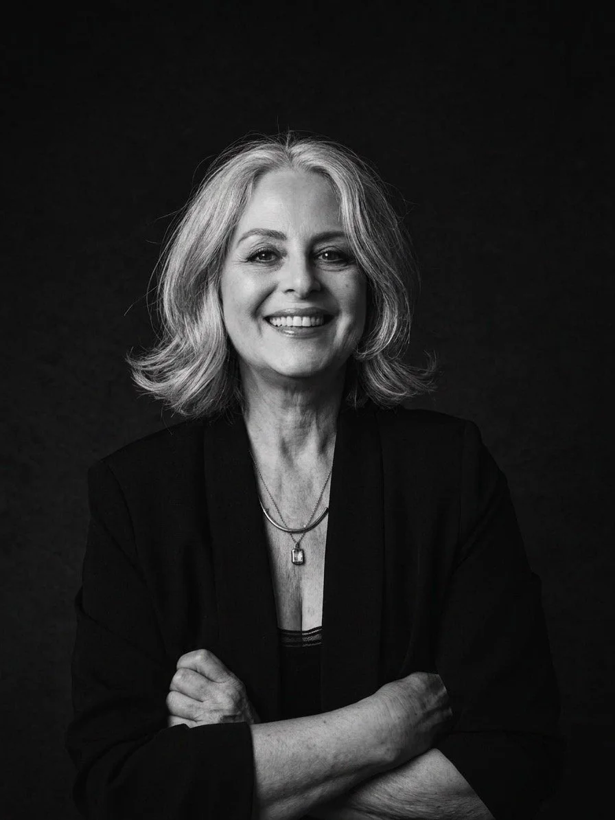 Black and white portrait of a smiling woman with shoulder-length wavy hair, wearing a dark blazer and a necklace, against a dark background.