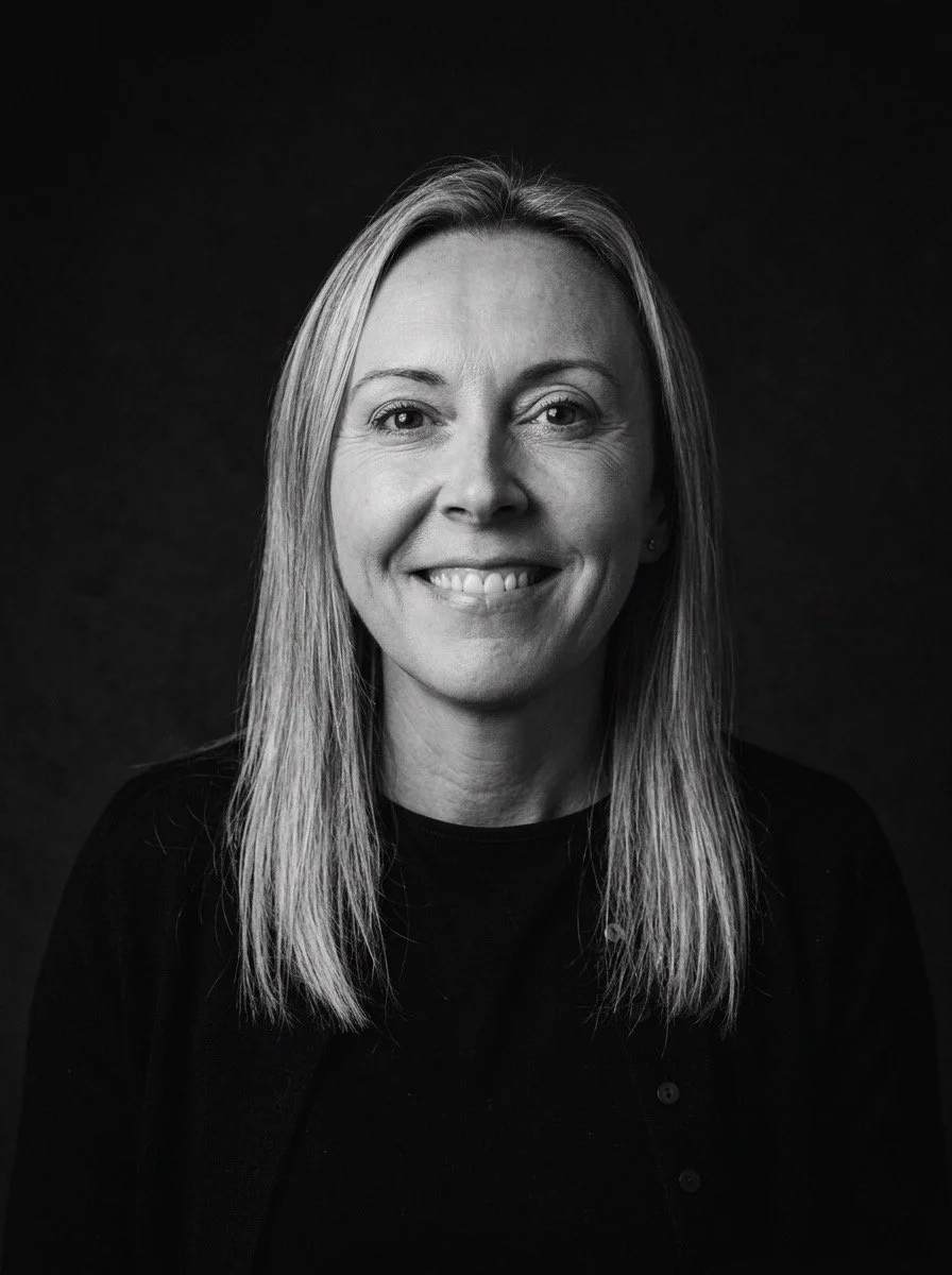 Black and white portrait of a smiling woman with shoulder-length hair, wearing a dark top, against a dark background.