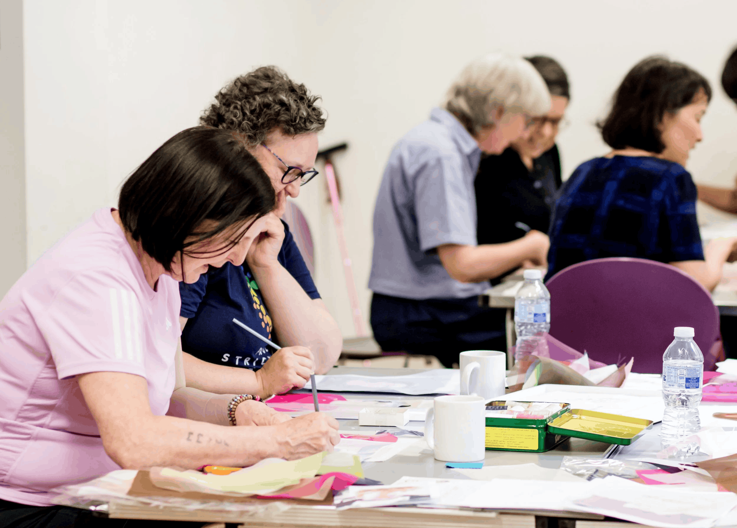 People sitting at a table working on arts and crafts, with various supplies like paper, pens, and water bottles visible.