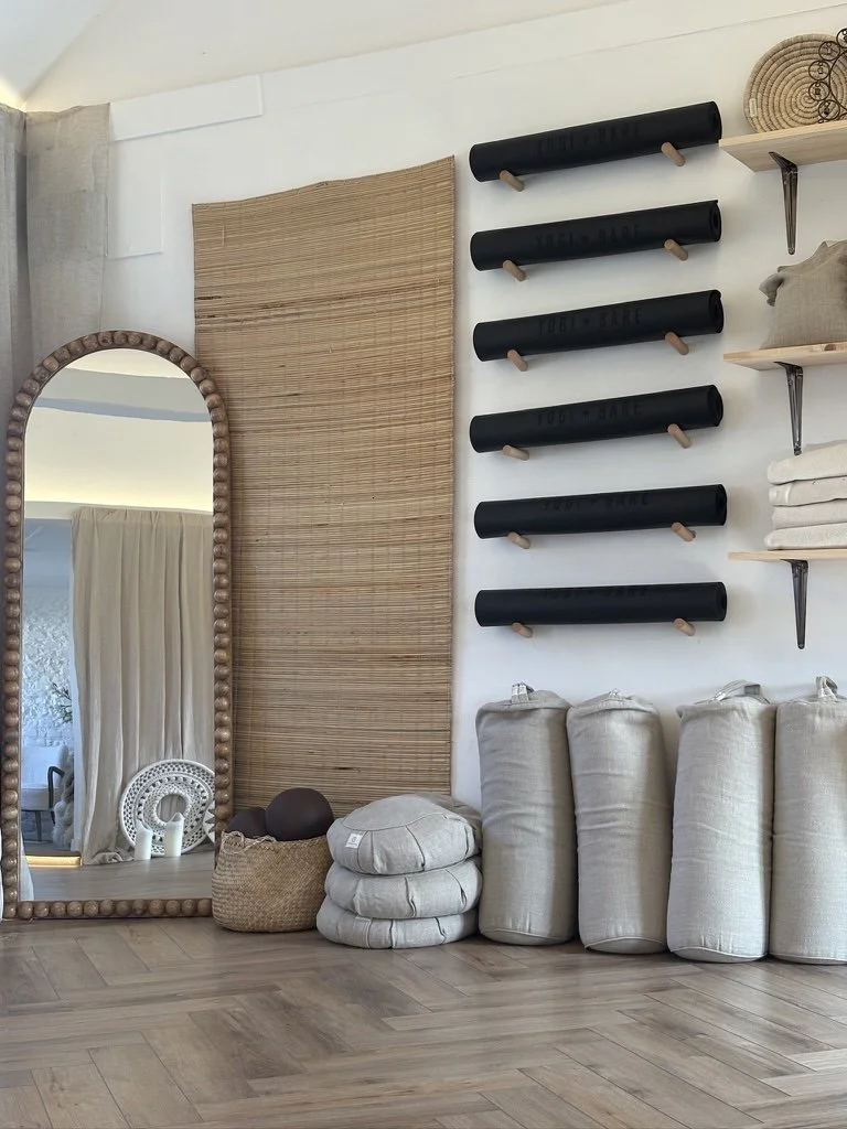 Decorative storage and accessories in a room with a large mirror, woven wall hanging, black Rolodex organizers, shelves with folded towels, fabric-covered bags, and cushions. Fully equipped luxury relaxing yoga studio in Sussex.
