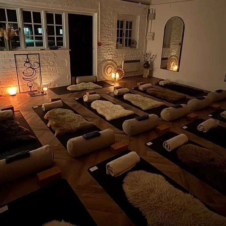 Yoga mats with bolster pillows and rolled towels arranged in a dimly lit, cozy room with candles, decorative walls, and a mirror. Yin yoga, restorative yoga, candlelit yoga, relaxing yoga Sussex.