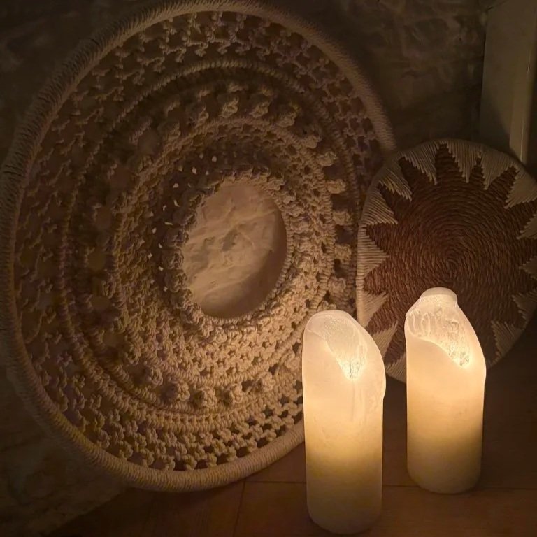Two glowing salt lamps placed on a wooden floor, with two decorative woven wall hangings with intricate patterns hanging behind them. Relaxing yoga and massage studio. 