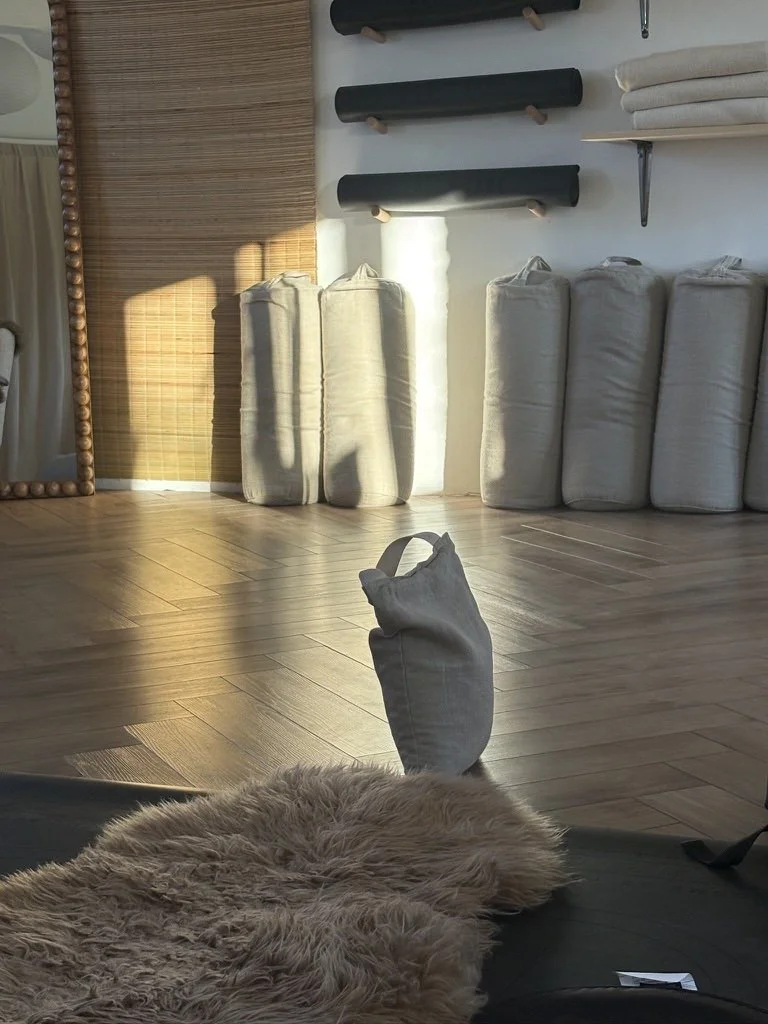 Indoor room with wooden floor, white walls, and shelves holding rolled mats and towels. A fluffy beige rug is on the floor, with a tote bag and part of a dark sofa visible in the foreground. Sunlight casts shadows on the floor.