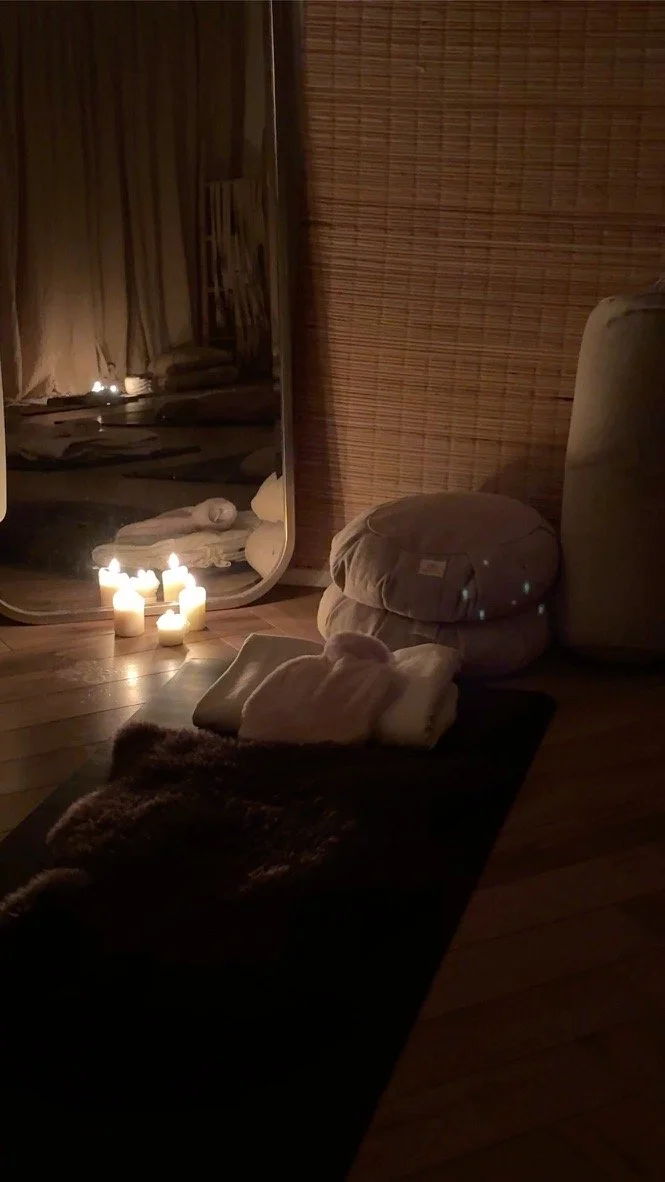 A cozy room with folded towels and linen stacked on a dark rug, lit by several candles on the wooden floor near a large mirror and a woven bamboo room divider.