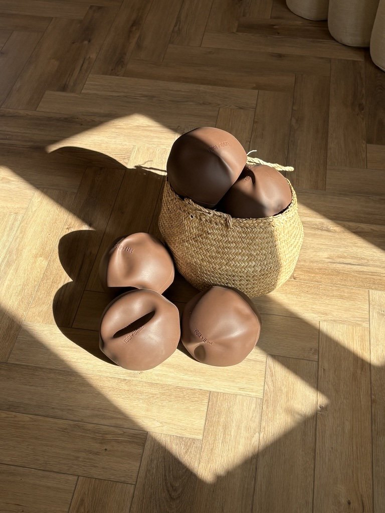 A wicker basket filled with plush toys resembling brown eggs with facial features, placed on a wooden floor in sunlight. Fully stocked yoga studio with luxury equipment. 