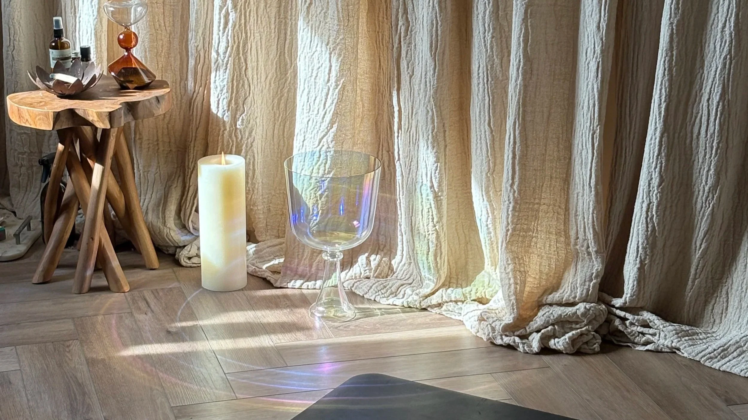 Decorative scene with a wooden side table holding glass bottles and a decorative bowl, a tall white candle, an empty glass, and a light-colored curtain with sunlight creating rainbow reflections on the floor in front of the curtain Sussex Yoga Studio