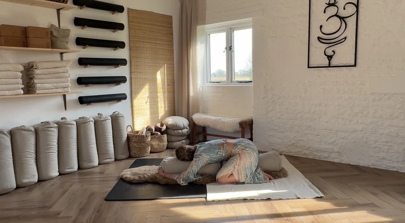A person practicing yoga in a peaceful, well-lit room with a wooden floor. The person is kneeling on a mat, resting their forehead on a bolster. The room has shelves with rolled towels and yoga props, a window letting in natural light, and wall art. The person is wearing light-colored, patterned yoga clothing.