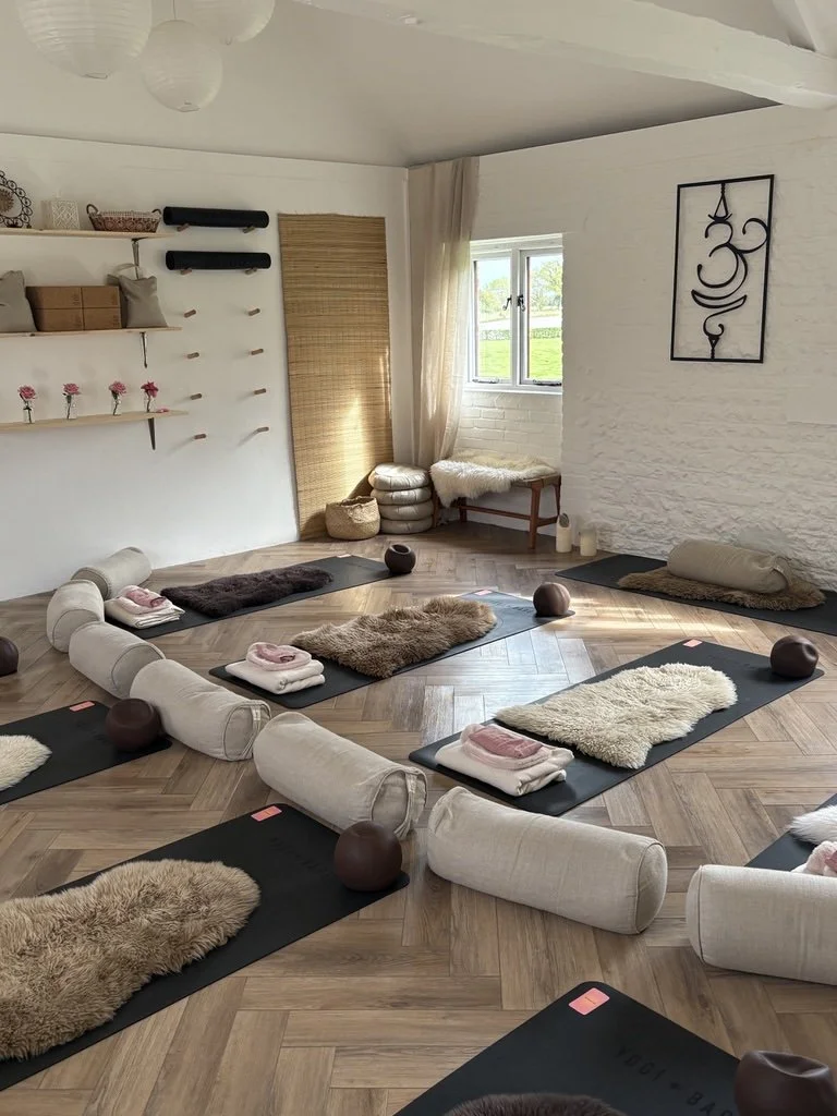 Yoga mats with blankets and meditation balls arranged in a circle in a well-lit, cozy room with white brick walls, a window with curtains, and decorative wall art.