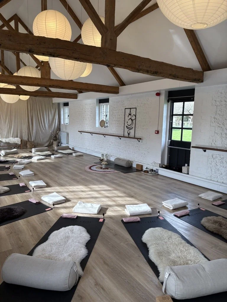 A spacious yoga studio with wooden beams on the ceiling, paper lantern lights, large windows, and a light-colored wooden floor. Yoga mats are laid out on the floor with blankets, towels, and bolsters, preparing for a class.